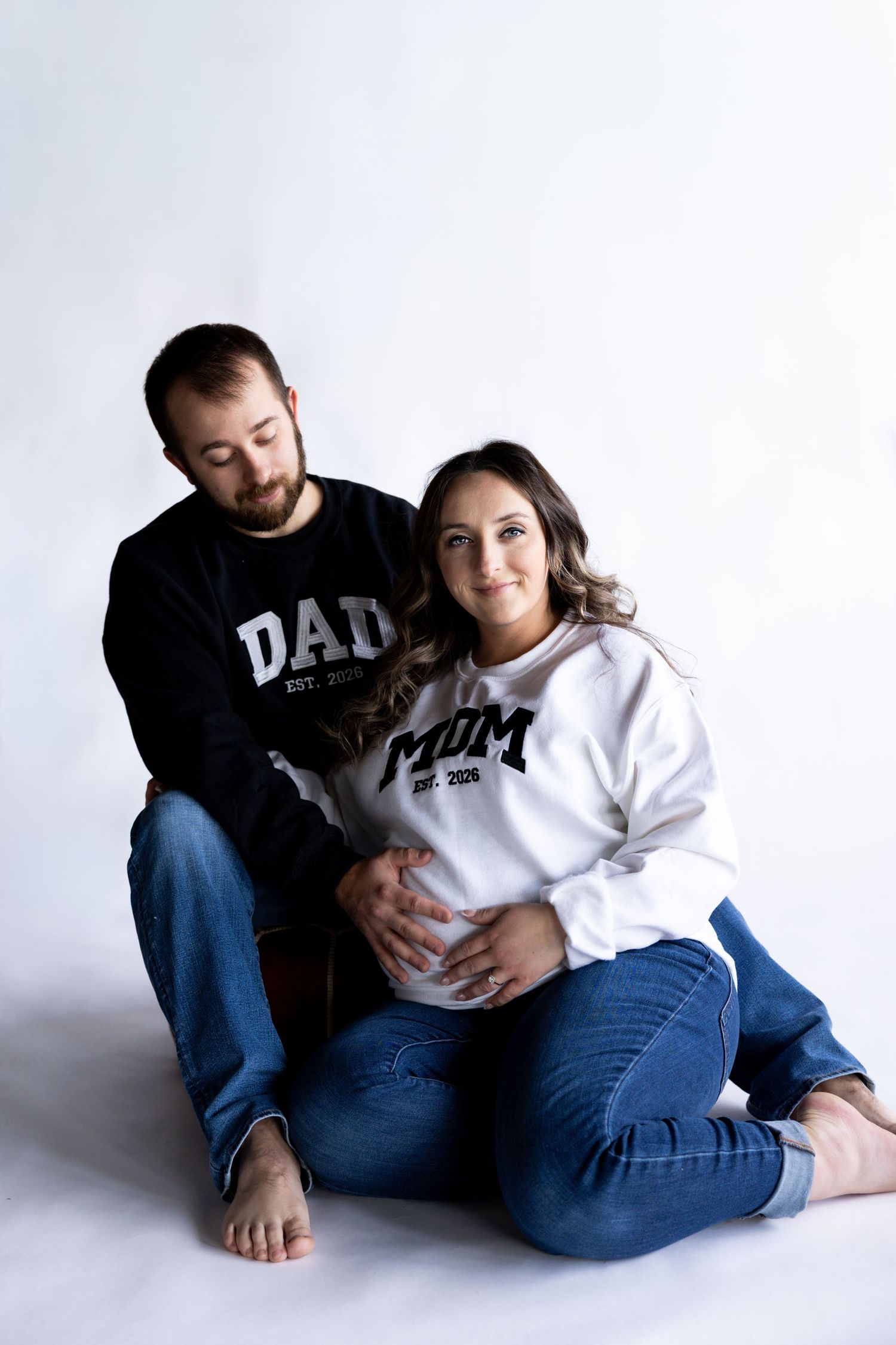 Casual maternity photo with couple in matching parent sweatshirts sitting on white floor.
