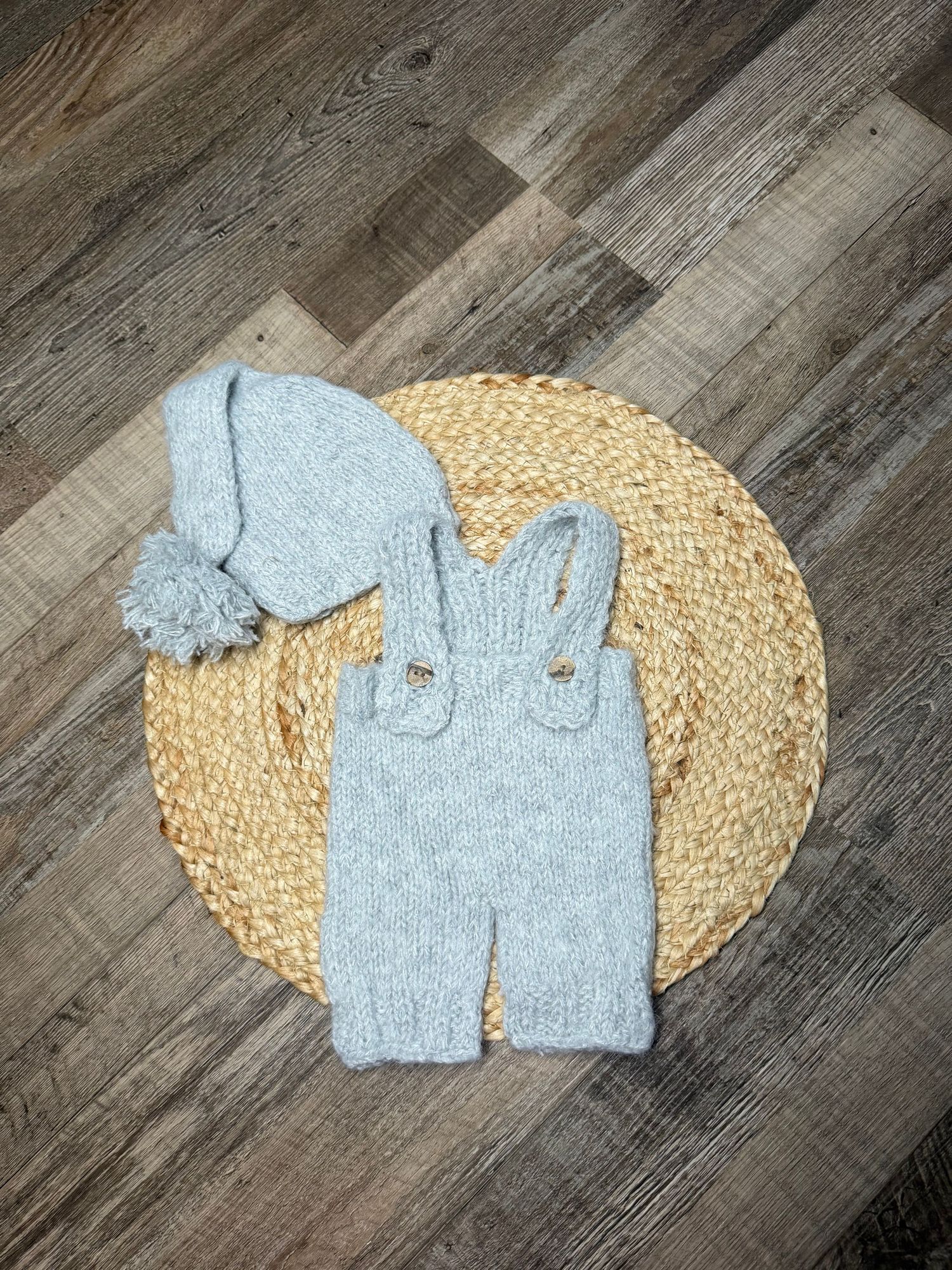 Light blue knitted baby outfit set with suspender pants and pom-pom hat displayed on woven placemat.