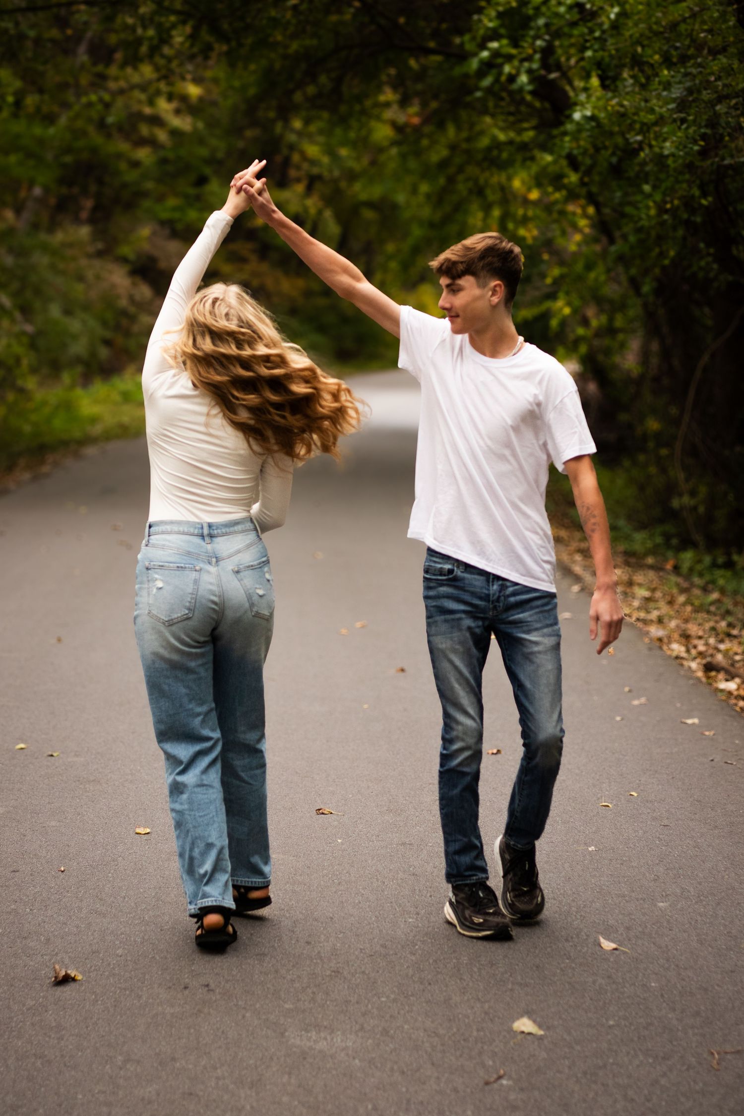 Two people in jeans and white shirts walk away on a path while holding hands and dancing together in a playful manner.