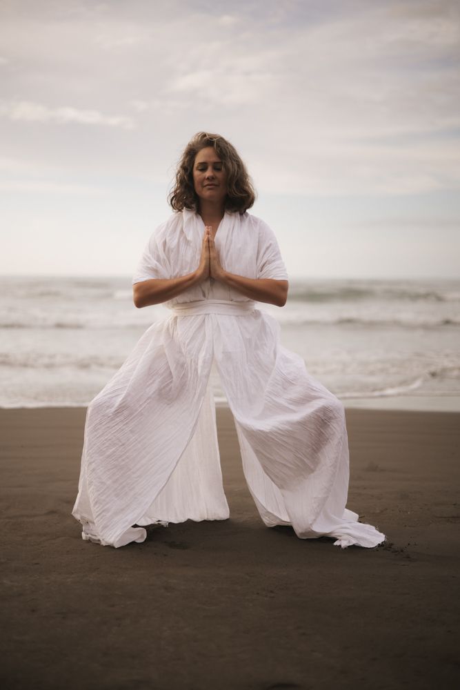 Woman standing on a black sand beach in a white kimono hands in prayer position in front of her chest.