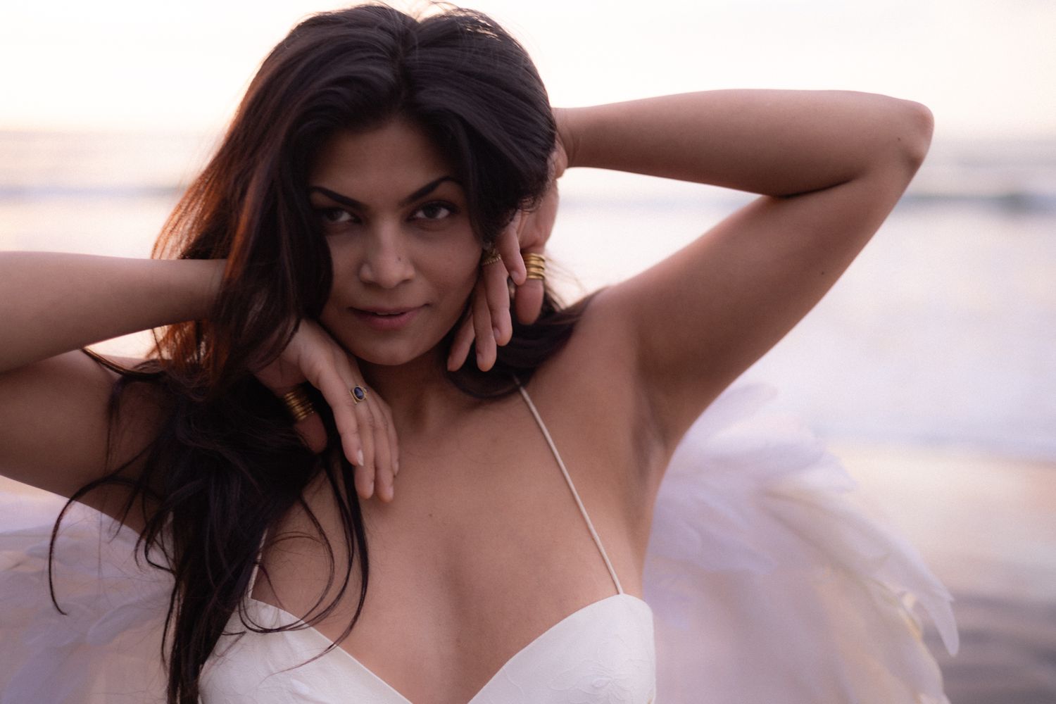 a close up of a woman on a back sand beach at sunset in a white dress with angel wings on her back looking seductive into the camera.