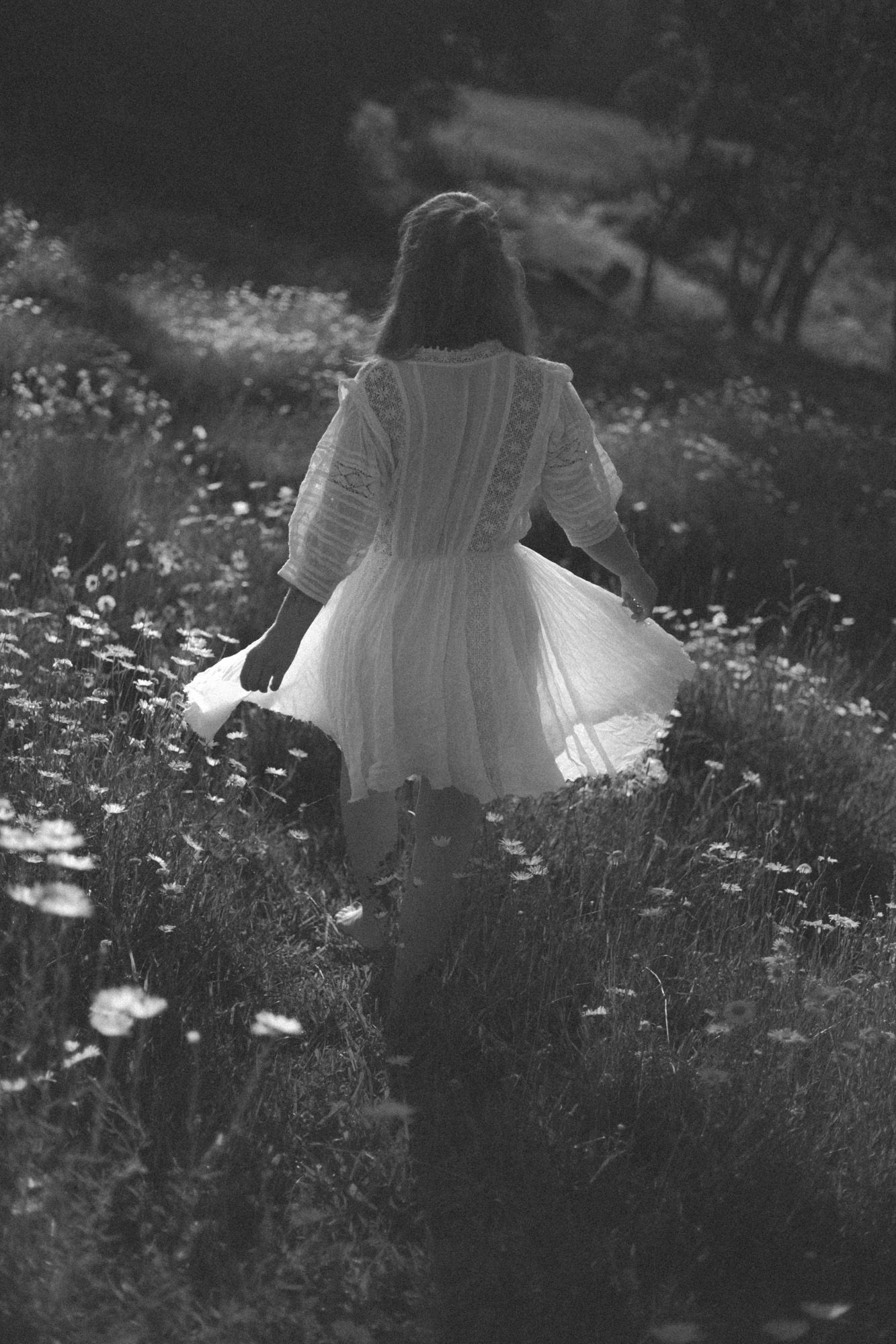 A woman walking away from the camera meandering through a field of knee high daisies in a white dress.