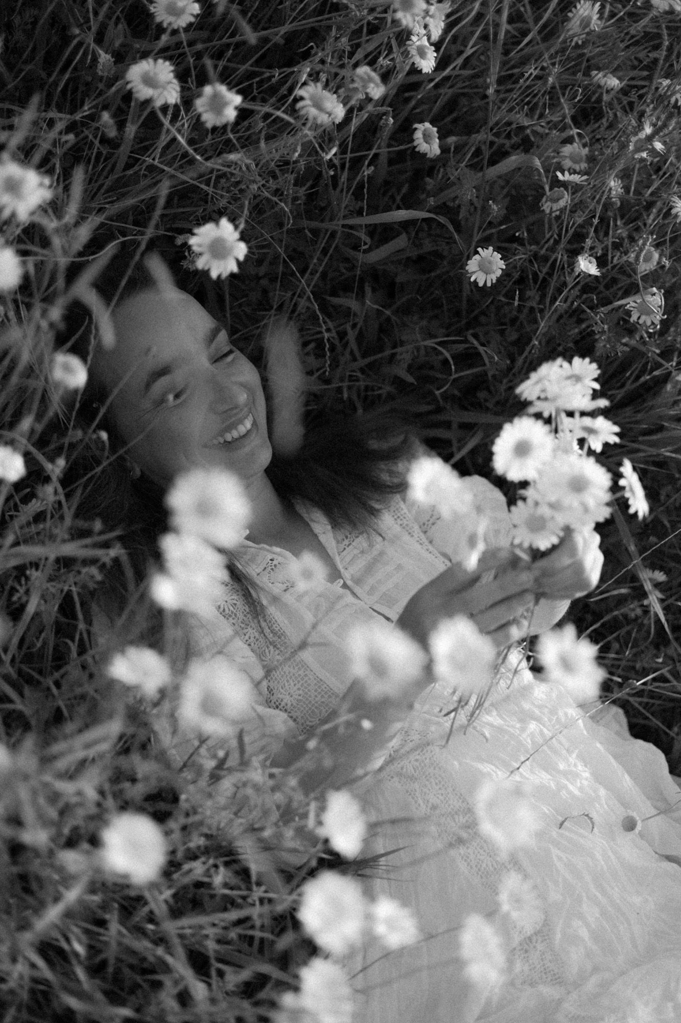 woman lying down in a field of daisies holding a bunch and laughing.
