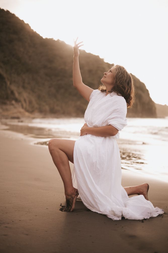 Woman sitting on a black sand beach in a white kimono on one knee looking and reaching up.