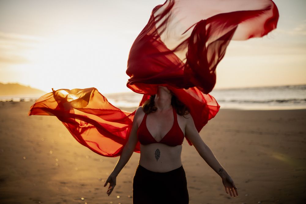 woman on the beach in red bikini, her head facing upward being covered by a big red silk like fabric that has been thrown up in the air