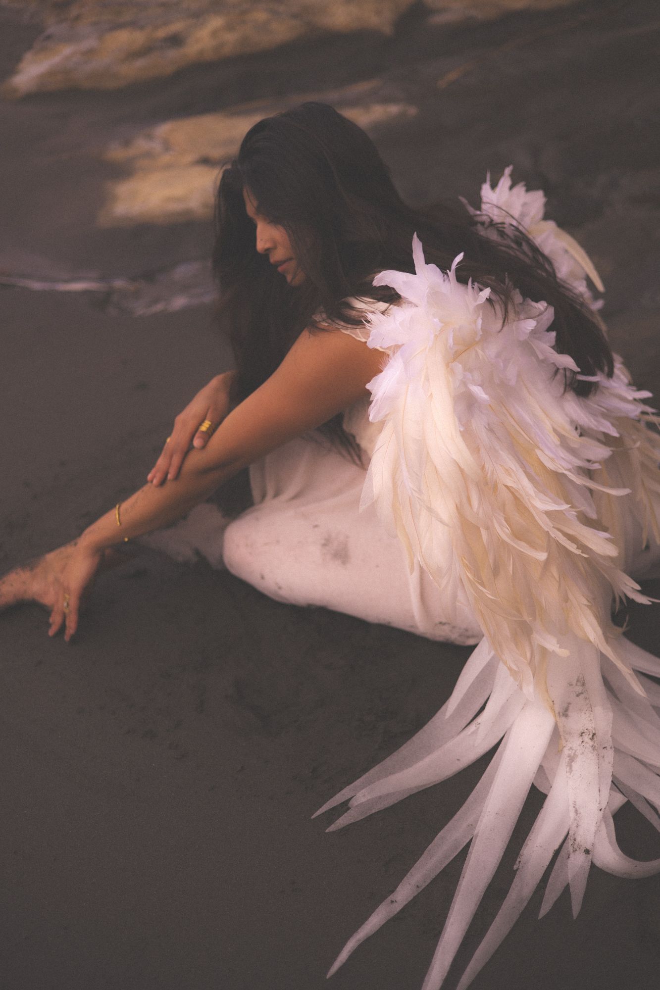 Woman on a back sand beach at sunset in a white dress with angel wings on her back sitting in the black sand getting dirty.