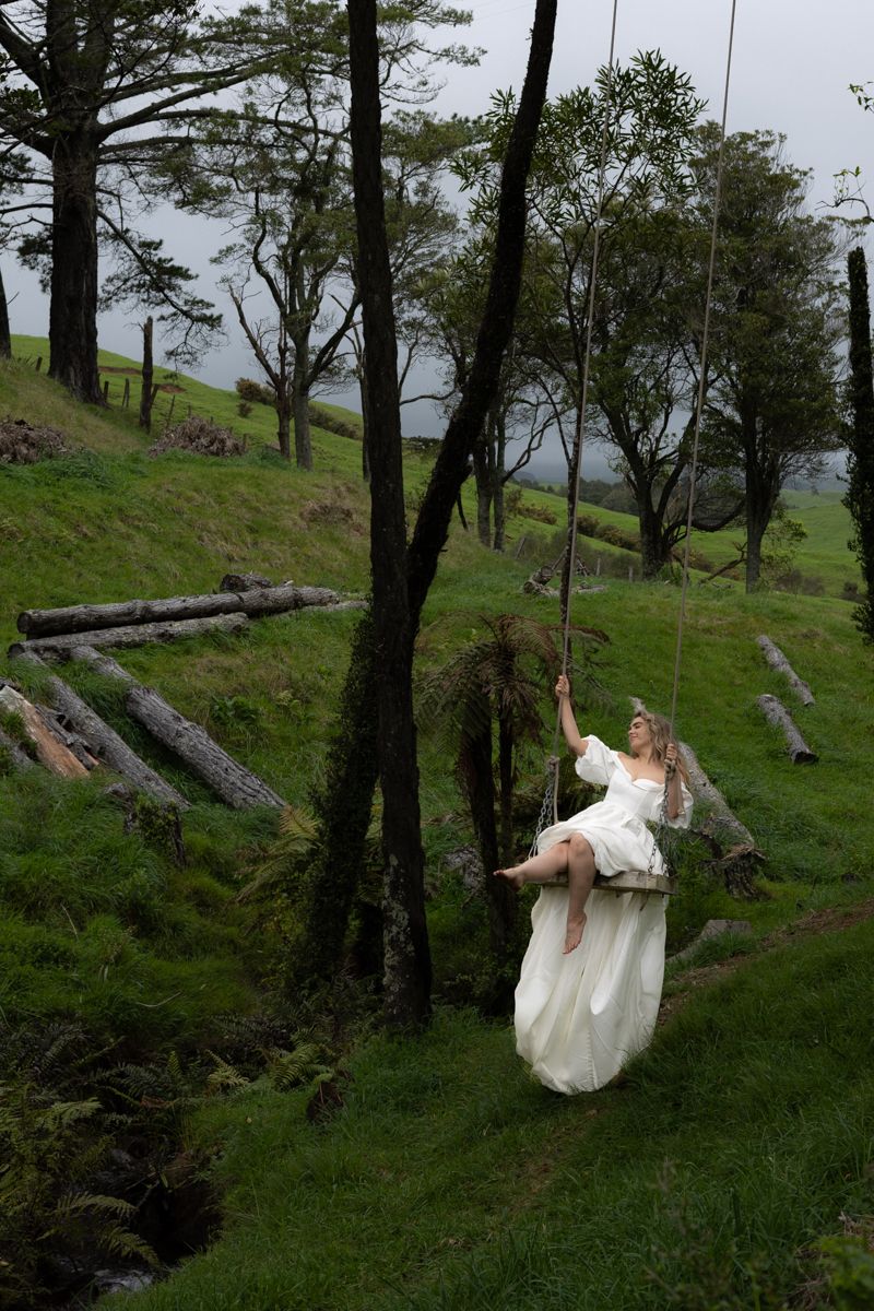 A woman in a long white wedding dress, by herself in a forest on a swing hanging from a tree branch, swinging happily.