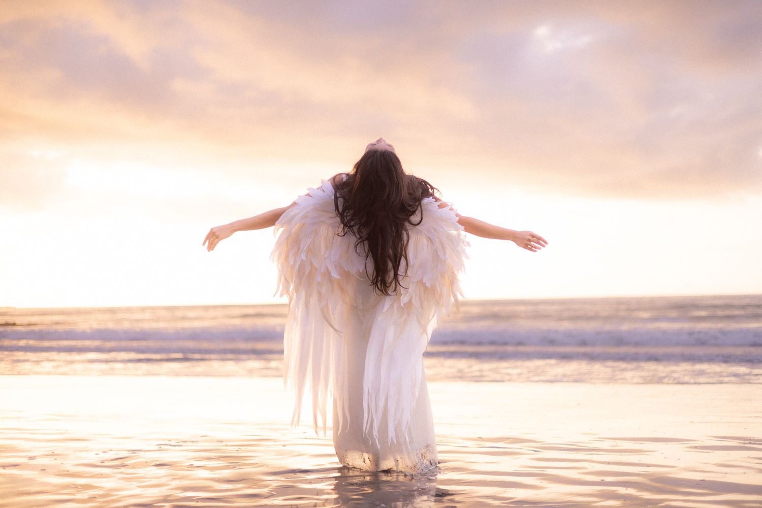 Woman on a back sand beach at sunset in a white dress with angel wings on her back with stretched out arms surrendering to the heavens above