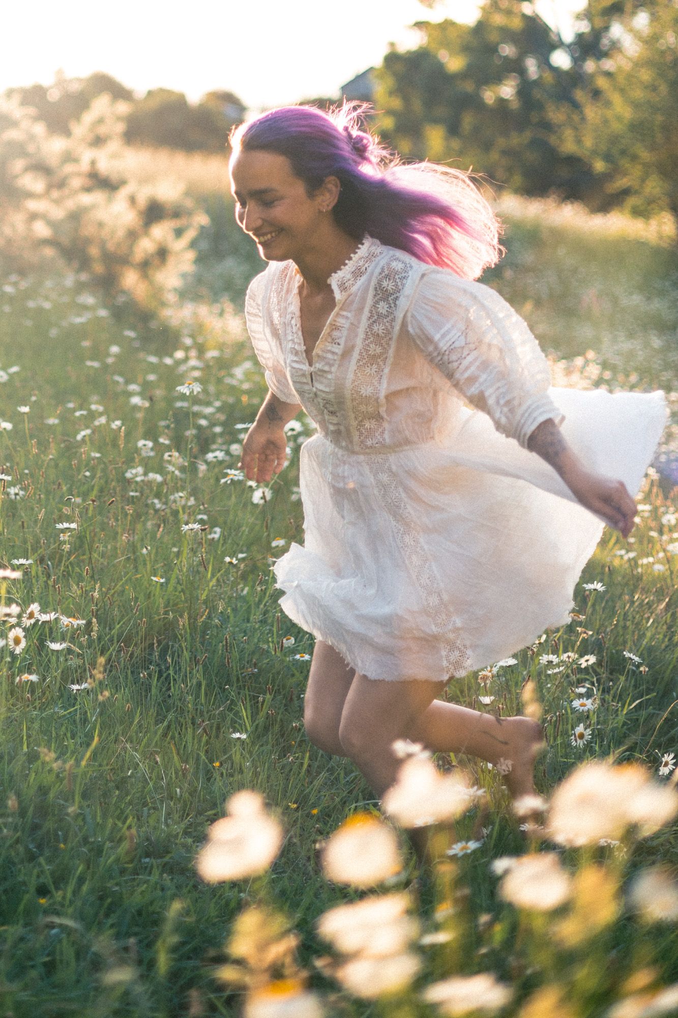 A woman running and laughing through a field of knee high daisies in a white dress at sunset.