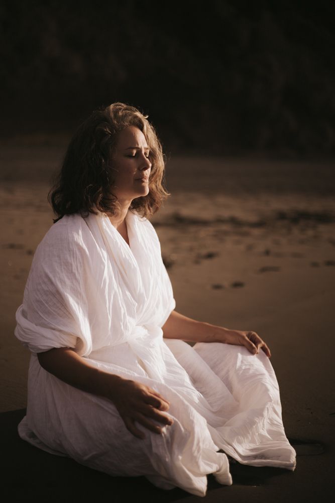 Woman sitting on a black sand beach in a white kimono with legs crossed meditating.