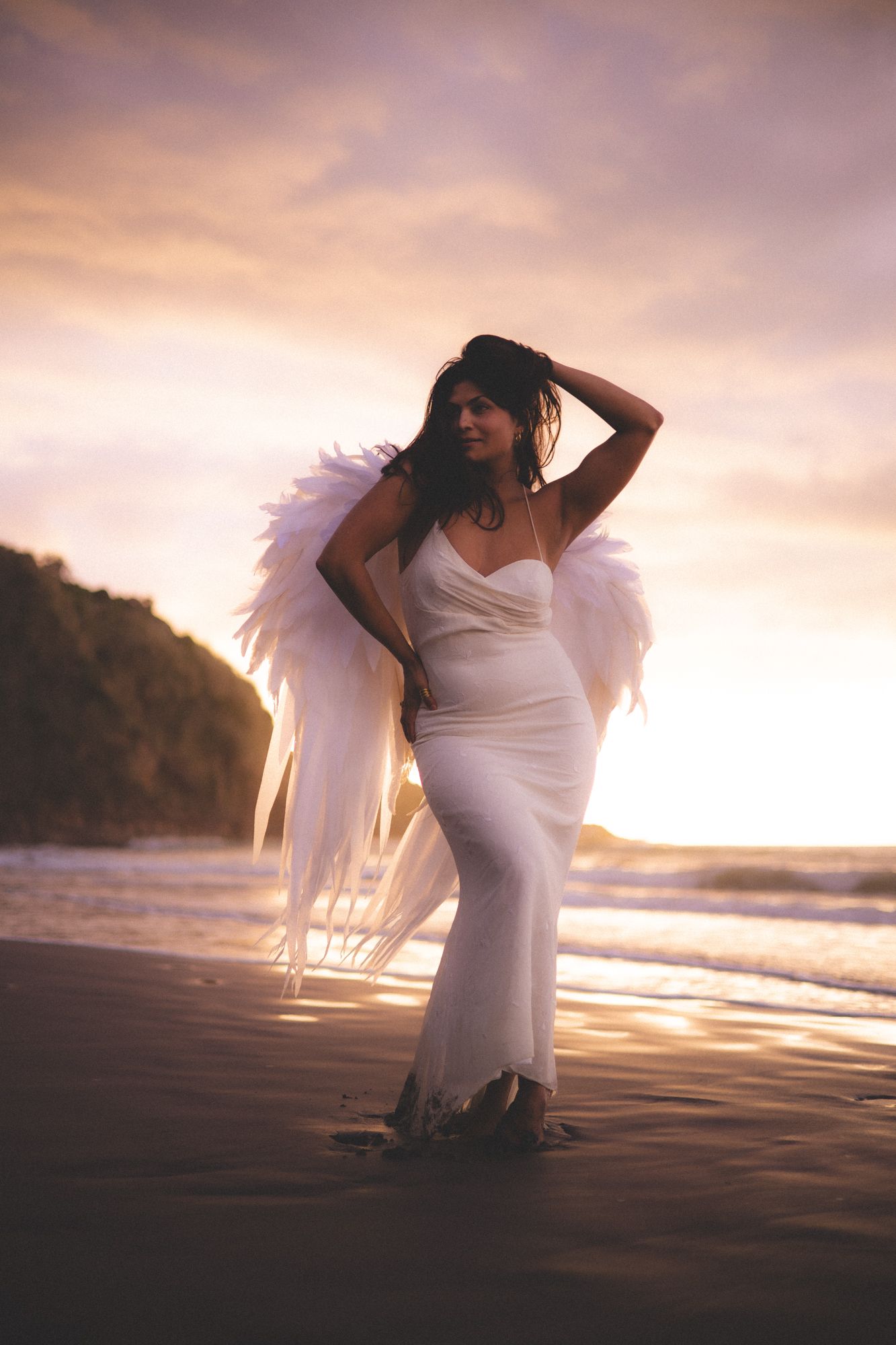 Woman on a back sand beach at sunset in a white dress with angel wings on her back