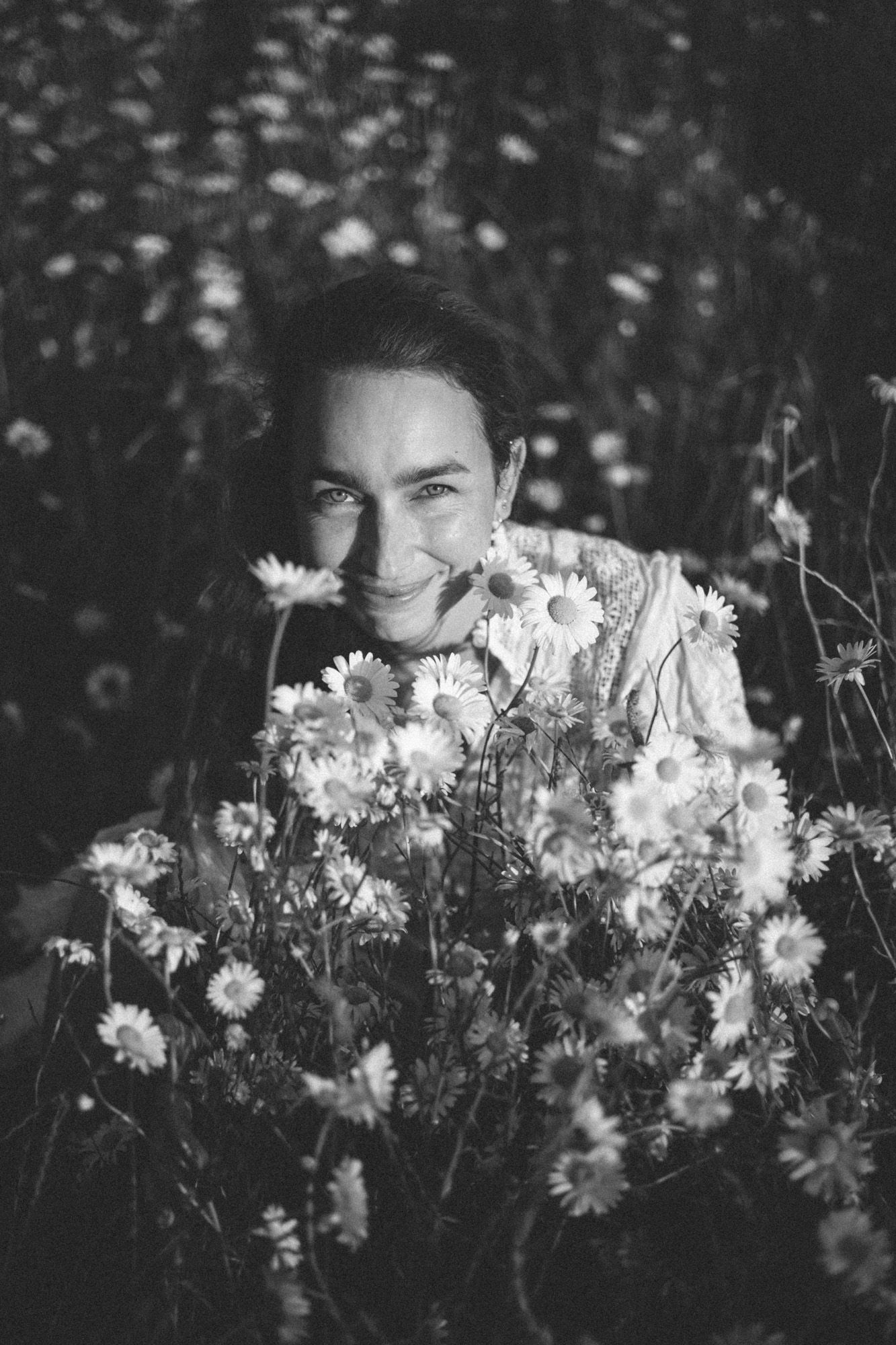 A woman sitting down in a field of knee high daisies gathering them in her arms making a massive bouquet looking up at the camera.