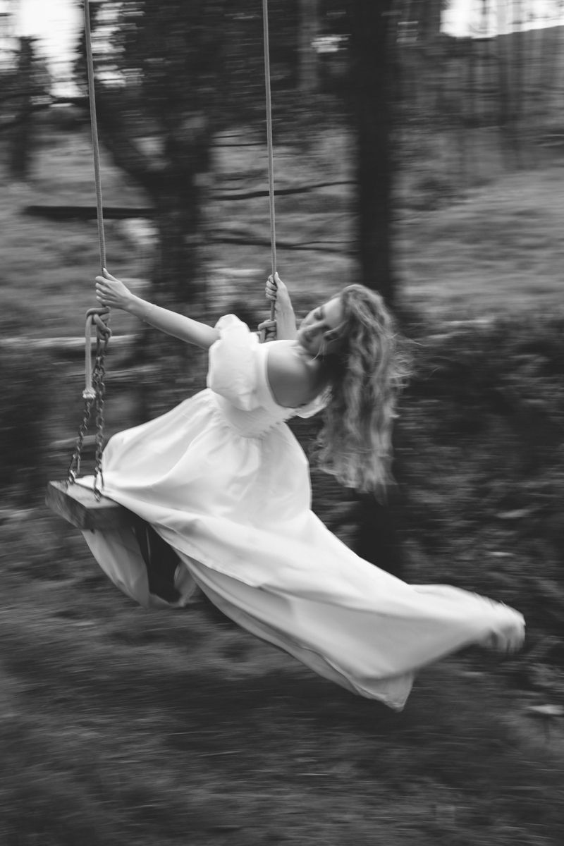 A woman in a long white wedding dress, by herself in a forest on a swing hanging from a tree branch, swinging happily.