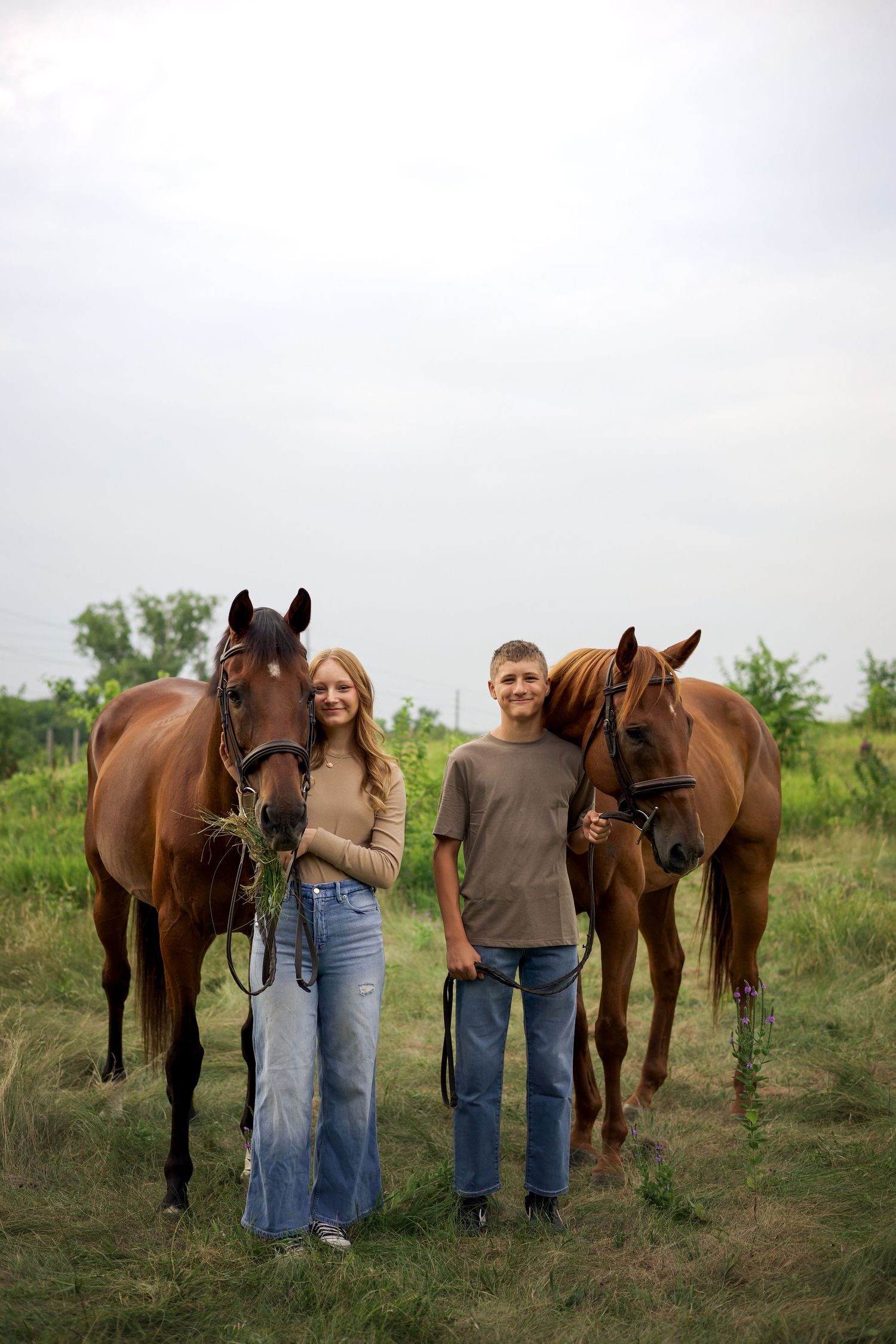 A young couple stands with two brown horses on a green field, holding reins and feeding hay on a cloudy day.