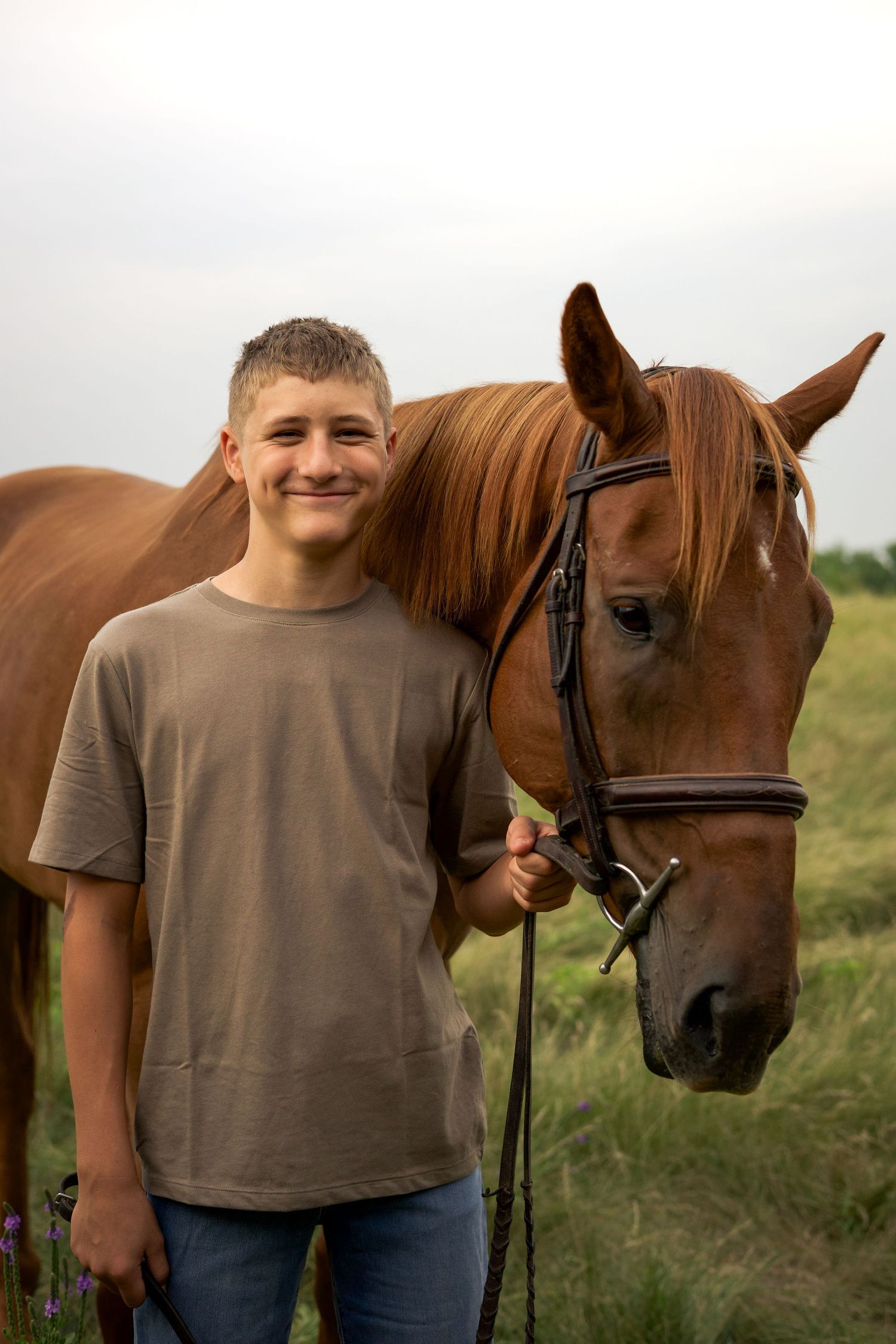 Smiling teenage boy standing next to a chestnut brown horse with a bridle in a green field.
