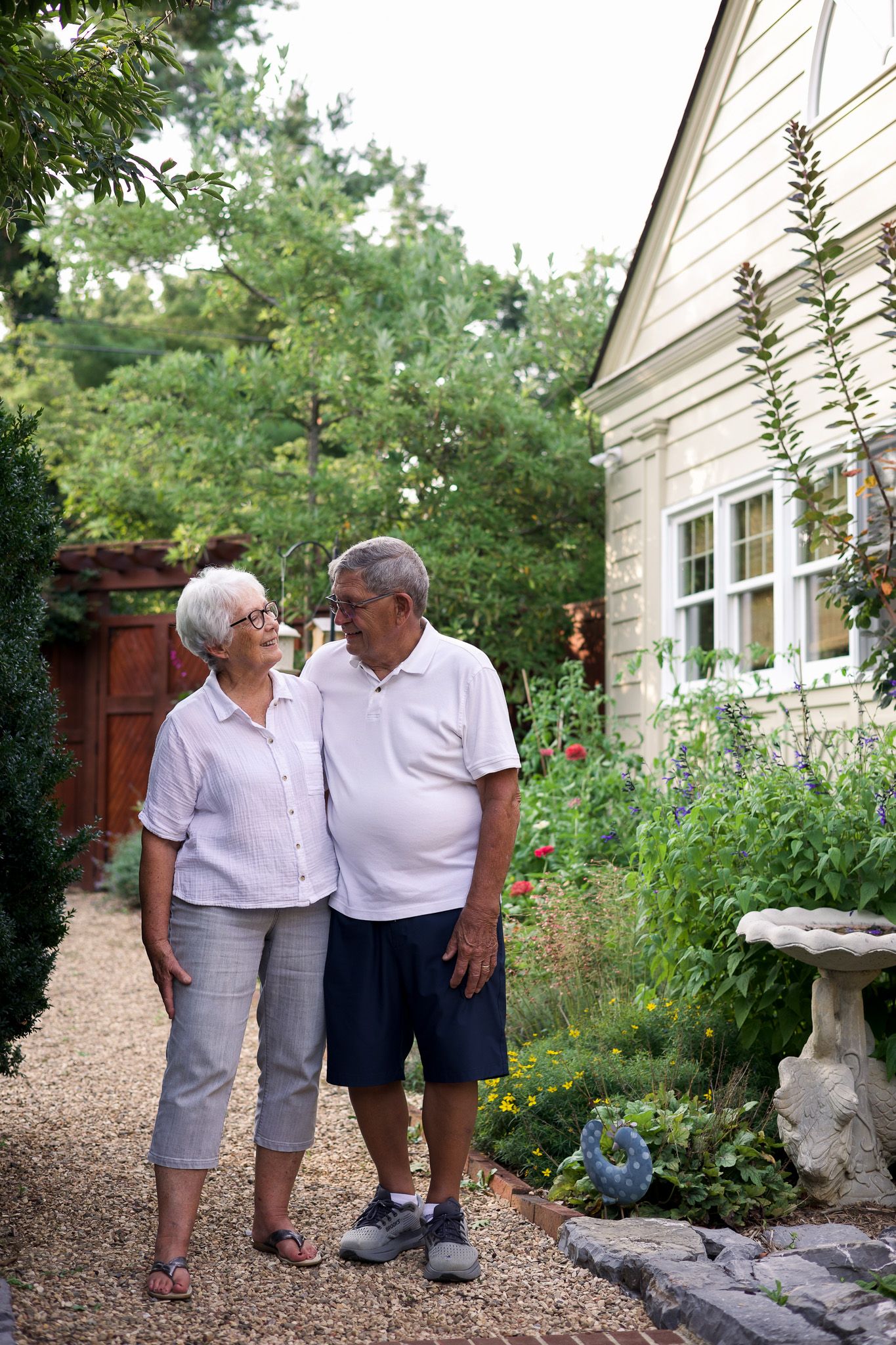 Senior couple standing together in a lush backyard garden beside their home, smiling at each other on a sunny day.