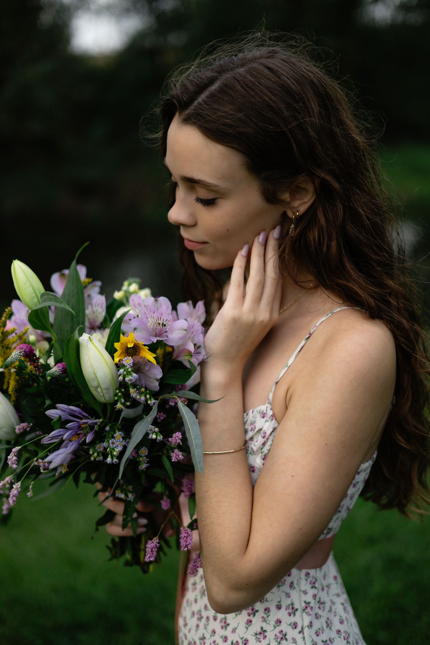 Young woman in floral dress holds colorful bouquet of lilies and wildflowers outdoors, gently touching her face.