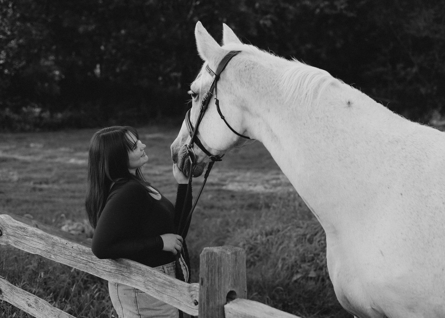 Black and white photo of a woman with long dark hair leaning on a wooden fence, gently touching the nose of a white bridled horse.