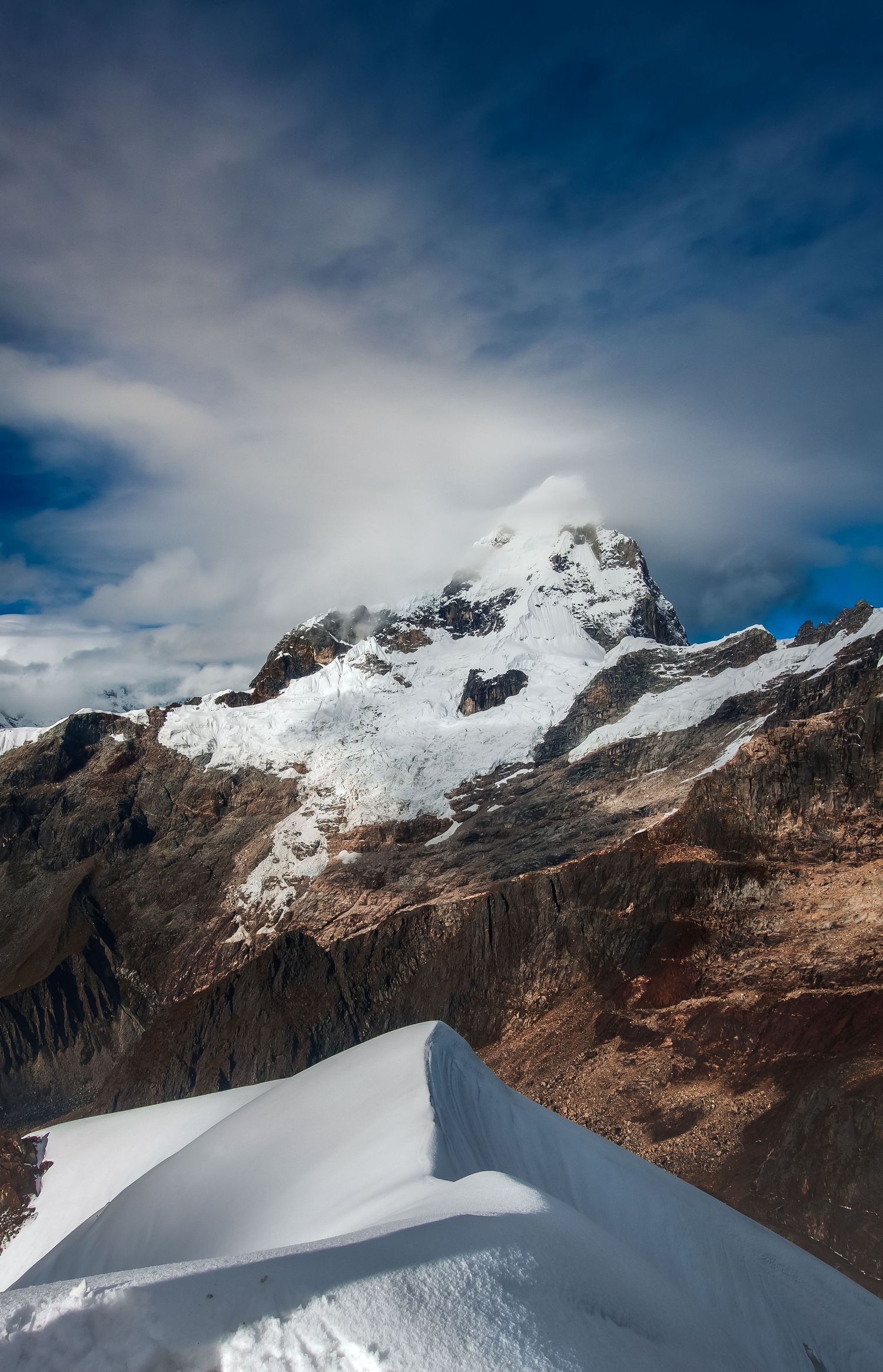 Snow-capped mountain peak with rocky terrain under dramatic blue sky with wispy clouds in alpine landscape.