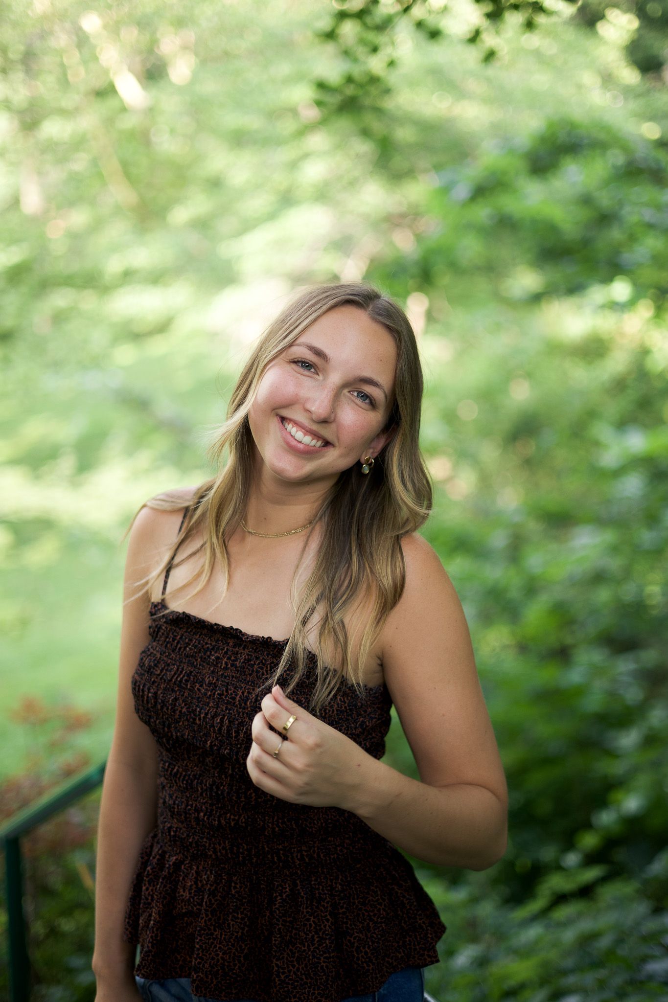 Smiling young woman with long blonde hair wearing a dark brown smocked top, posing outdoors among lush green trees.