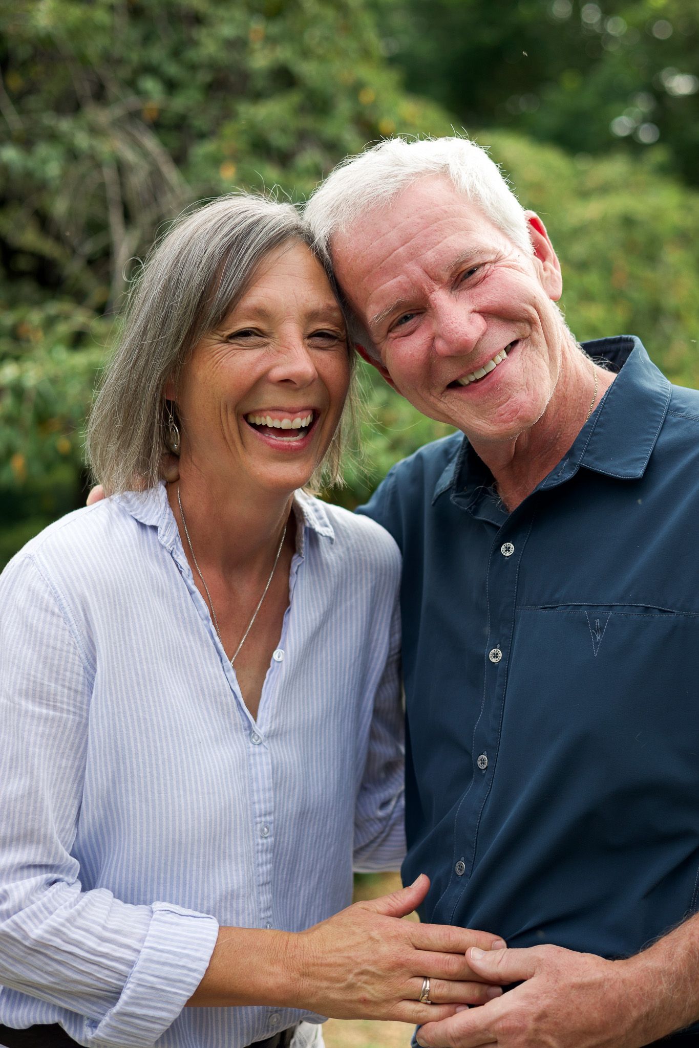 Happy senior couple laughing and embracing outdoors, with lush green trees in the background.
