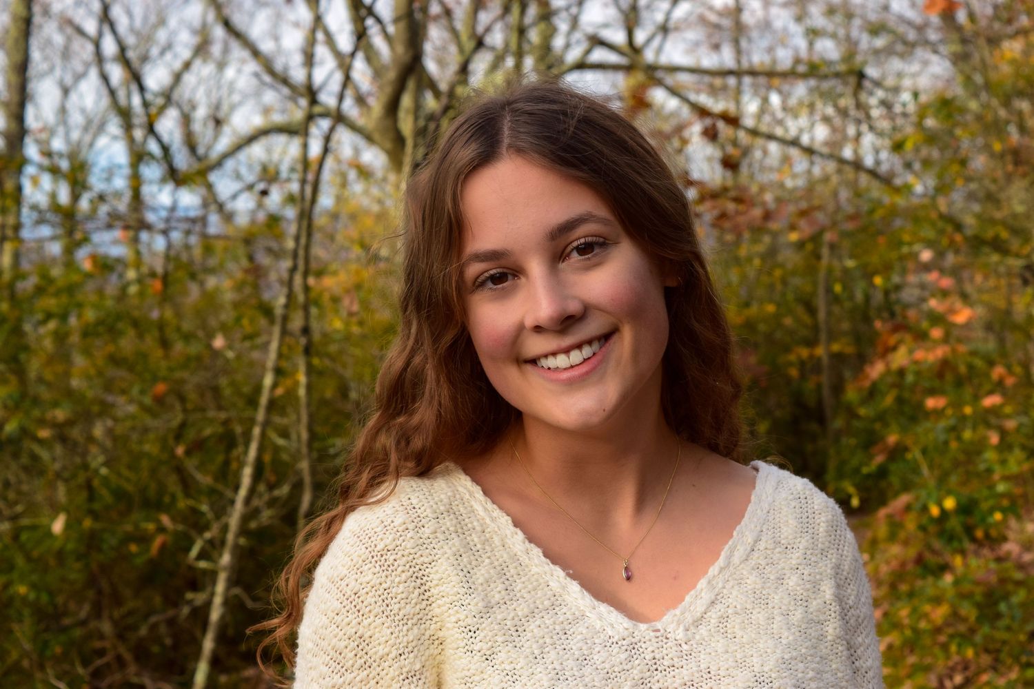 Smiling young woman with brown hair wearing a cream knit sweater outdoors among autumn trees.