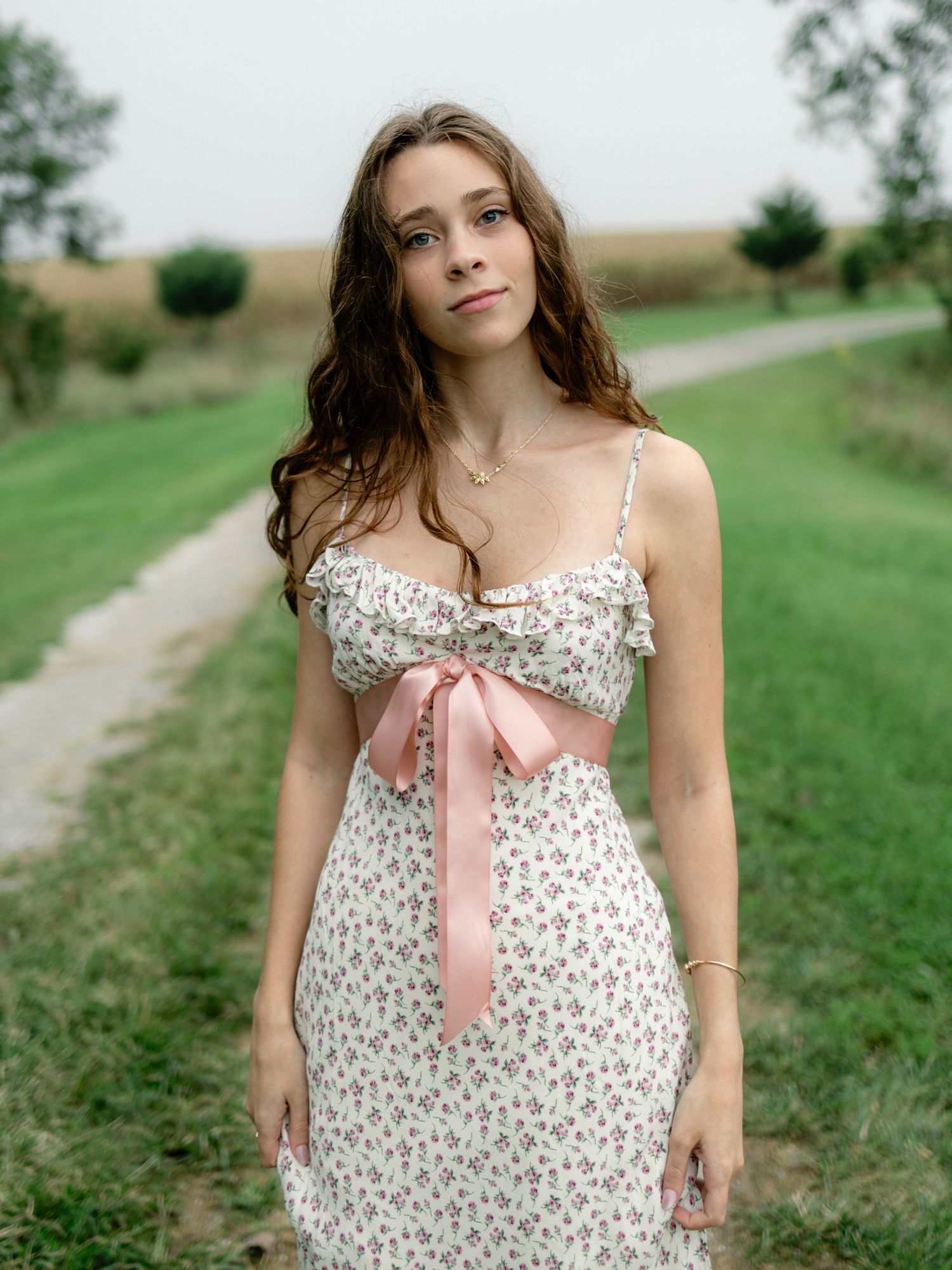 Young woman in floral sundress with pink bow poses on a scenic green country path.