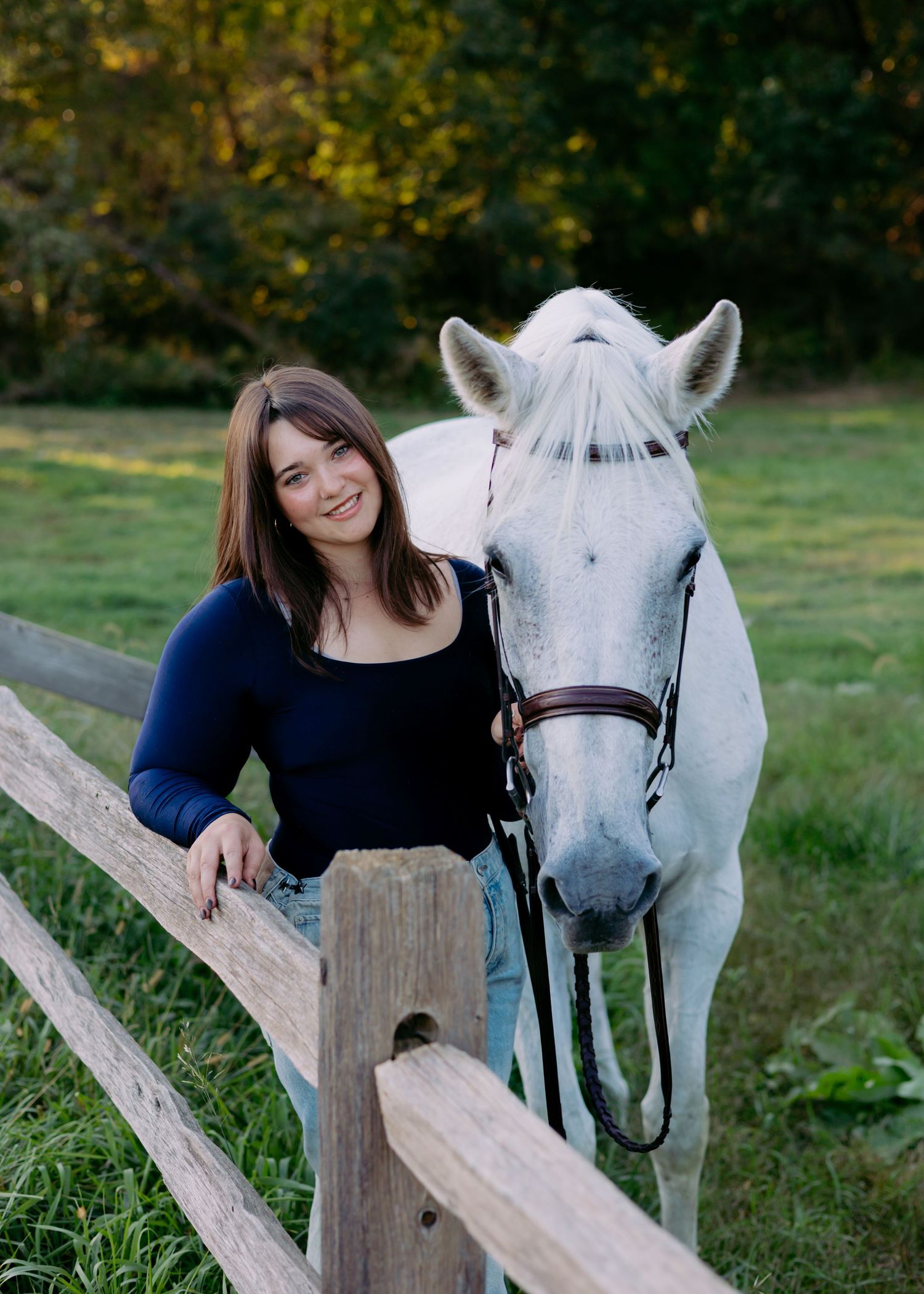 Smiling woman in a navy top leans on a wooden split-rail fence beside a white horse in a bridle on a sunlit grassy pasture.