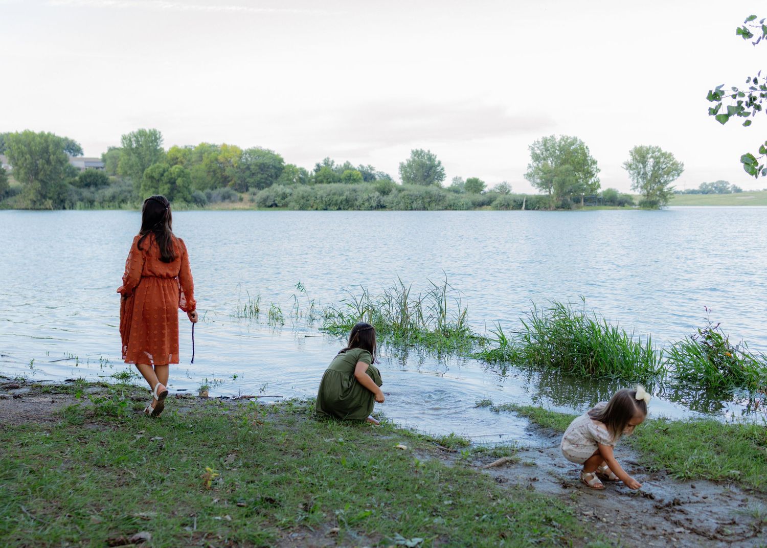 Three people stand and crouch along a grassy lake shore at dusk, one in an orange dress, with calm water and distant trees behind them.