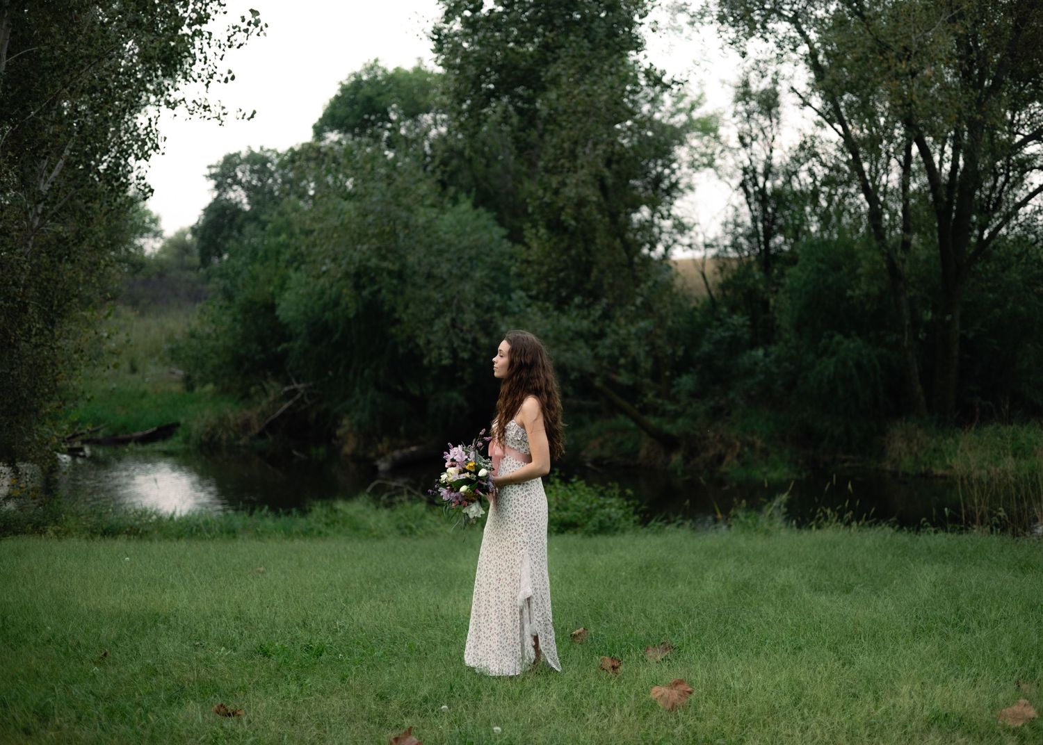Bride in lace gown holding bouquet stands in lush green meadow near creek surrounded by trees.