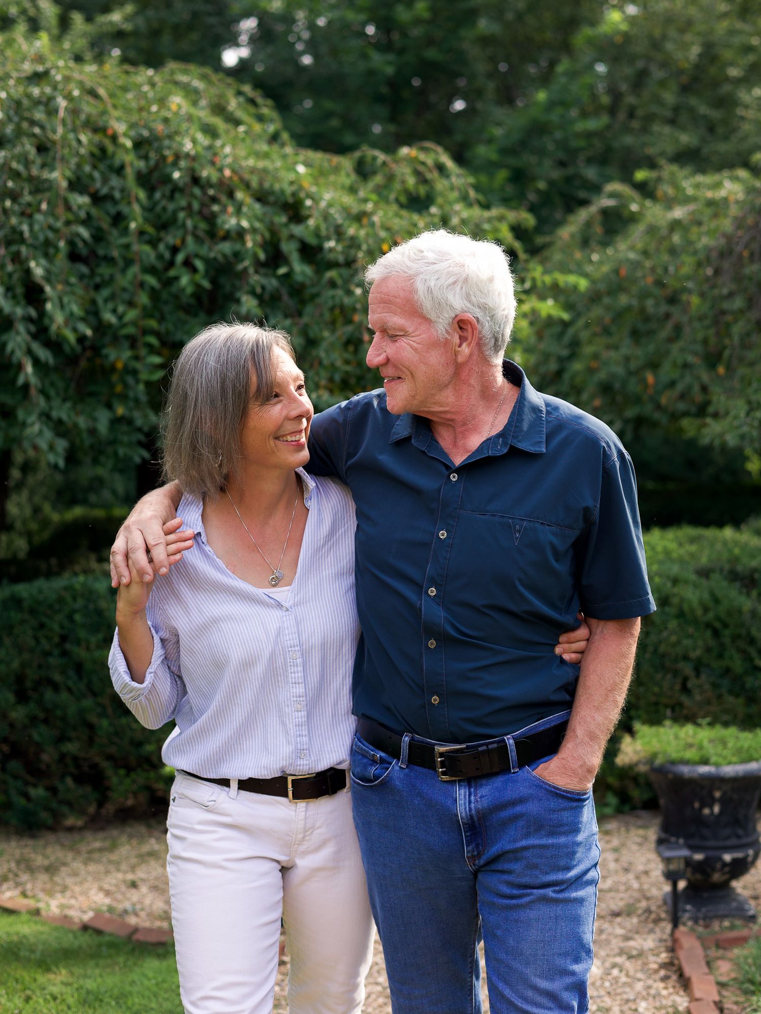 Older couple smiling and walking together in a lush garden, man in blue shirt and jeans, woman in white pants.