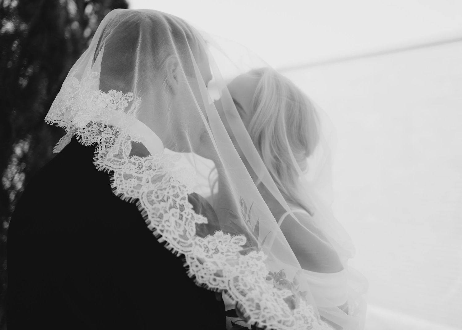 Black and white photo of a bride and groom embracing under a delicate lace-trimmed wedding veil.