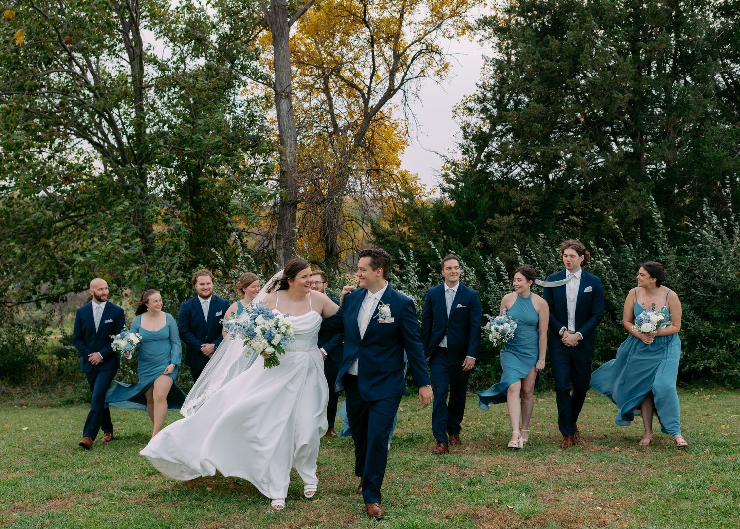 Bride in white gown and groom in navy suit walk laughing across a grassy lawn with their wedding party in blue dresses and suits.