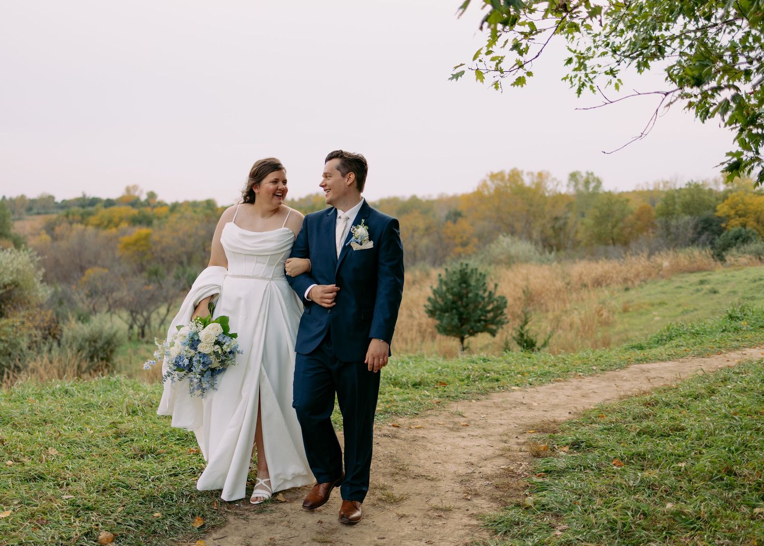Bride in a white gown holding a bouquet walks hand in hand with a groom in a navy suit along a dirt path through autumn fields.