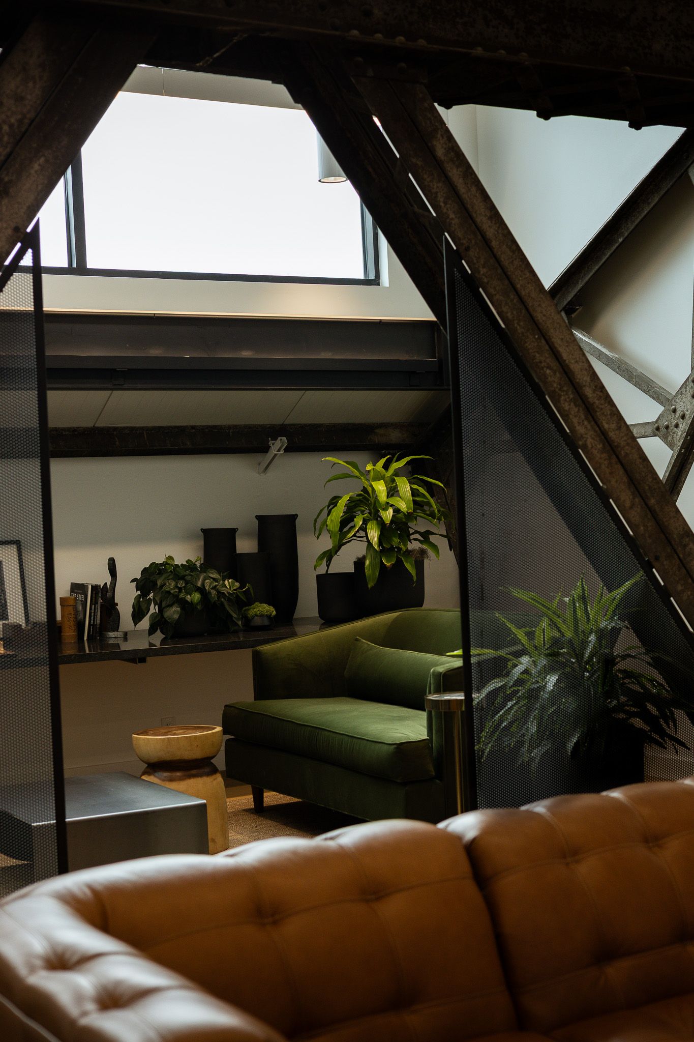 Industrial loft living room with leather sofa, green velvet armchair, tropical plants, and exposed wooden beams under skylights.
