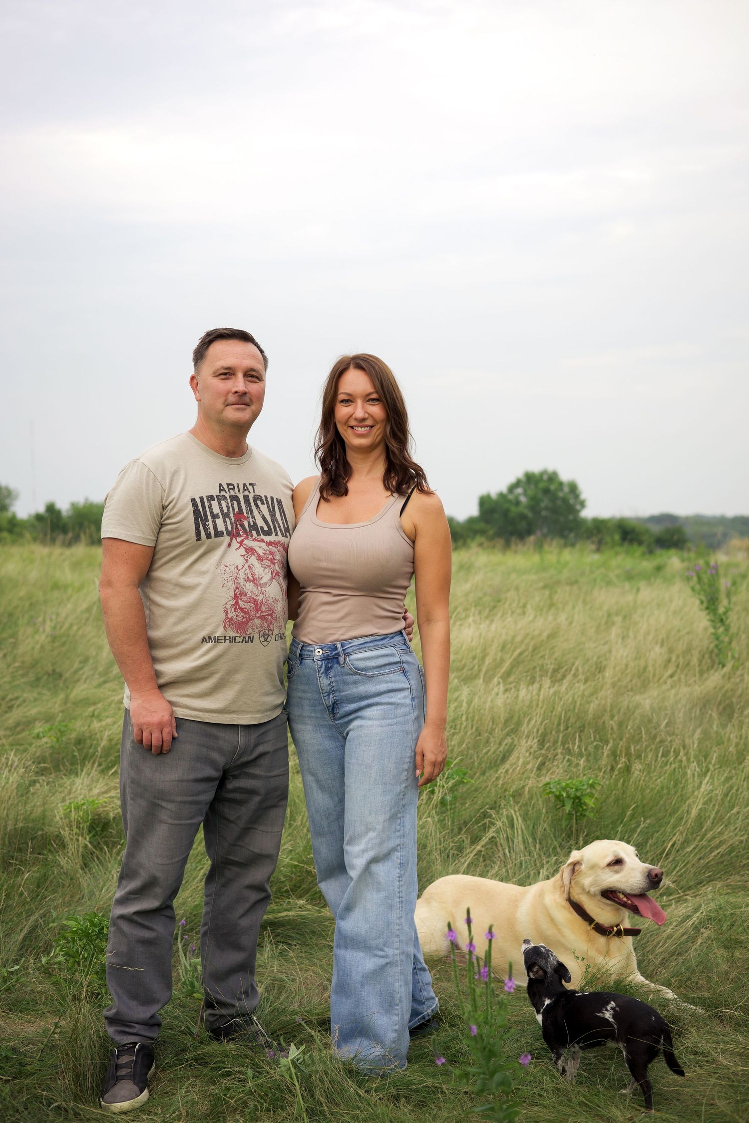 A couple smiles outdoors in a green field with their yellow Lab and small black dog on a cloudy day.