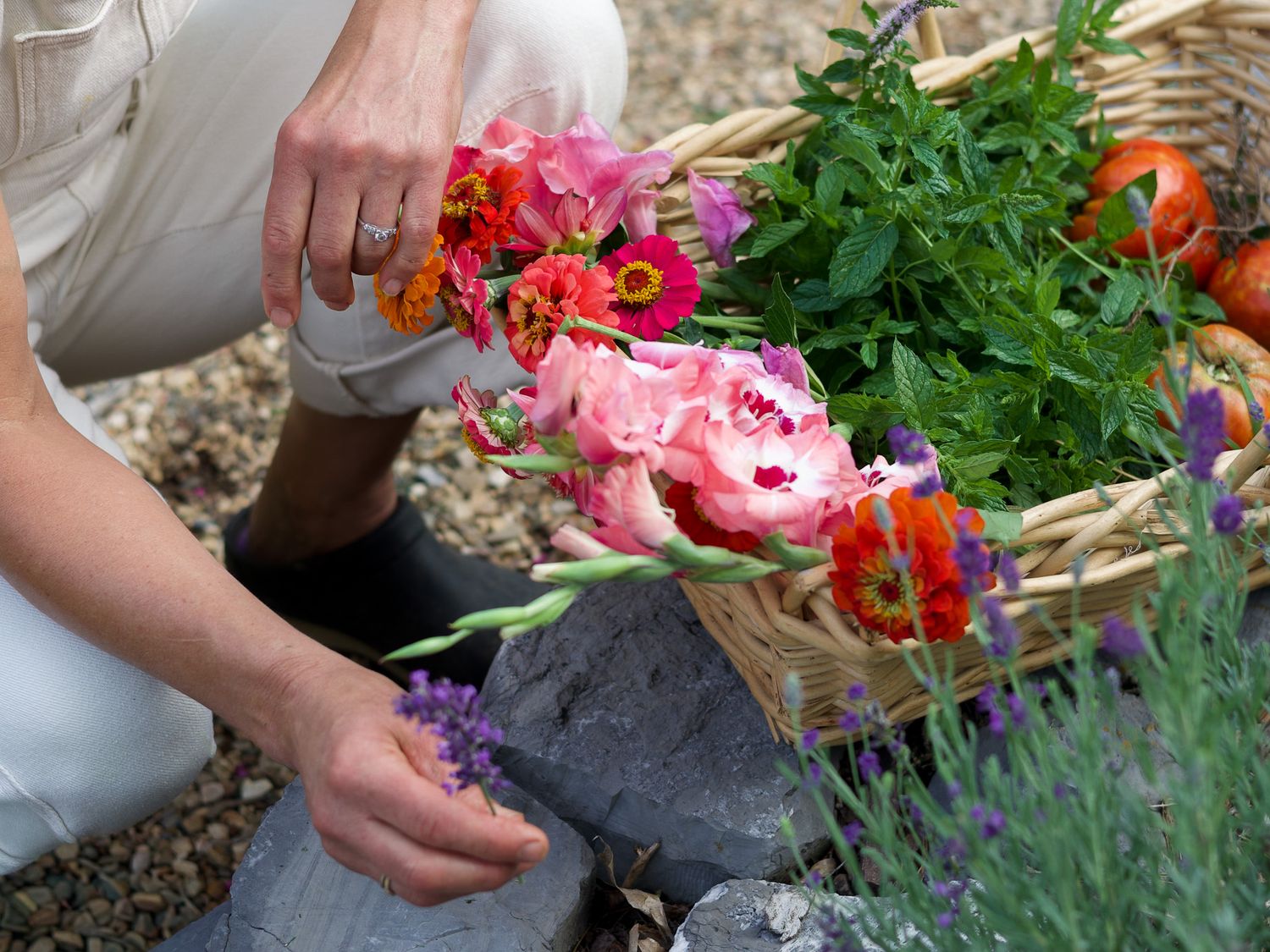 Person kneeling in garden arranging colorful flowers beside a wicker basket filled with fresh blooms and vegetables.