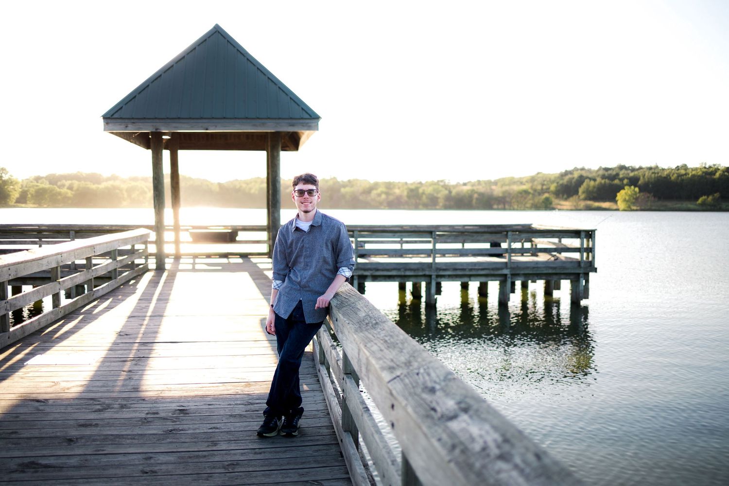 Man in a blue button-up shirt and glasses leans on a wooden railing of a lakeside pier with a gazebo at sunset.