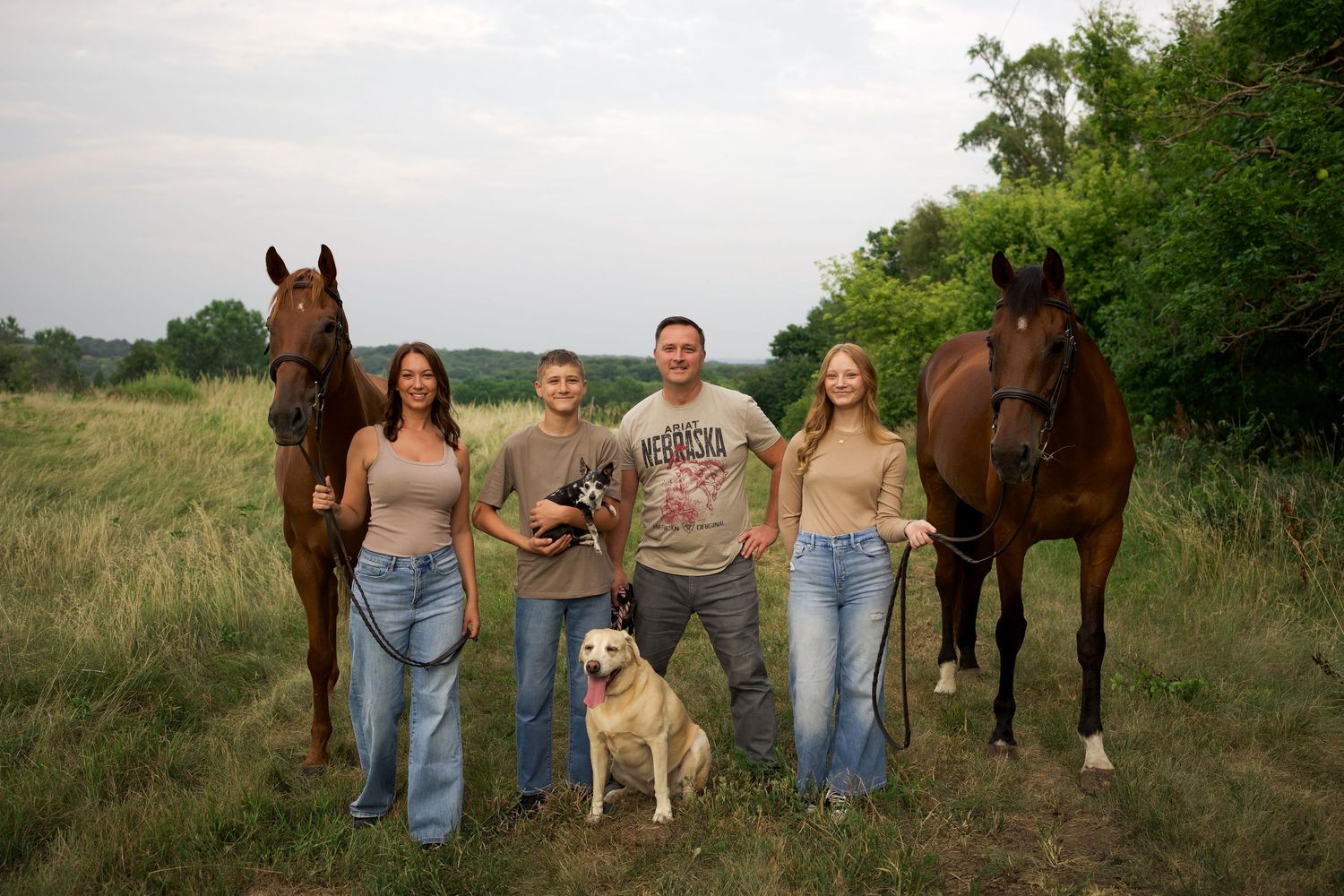 Family of four poses with two brown horses and a yellow lab in a lush green field.