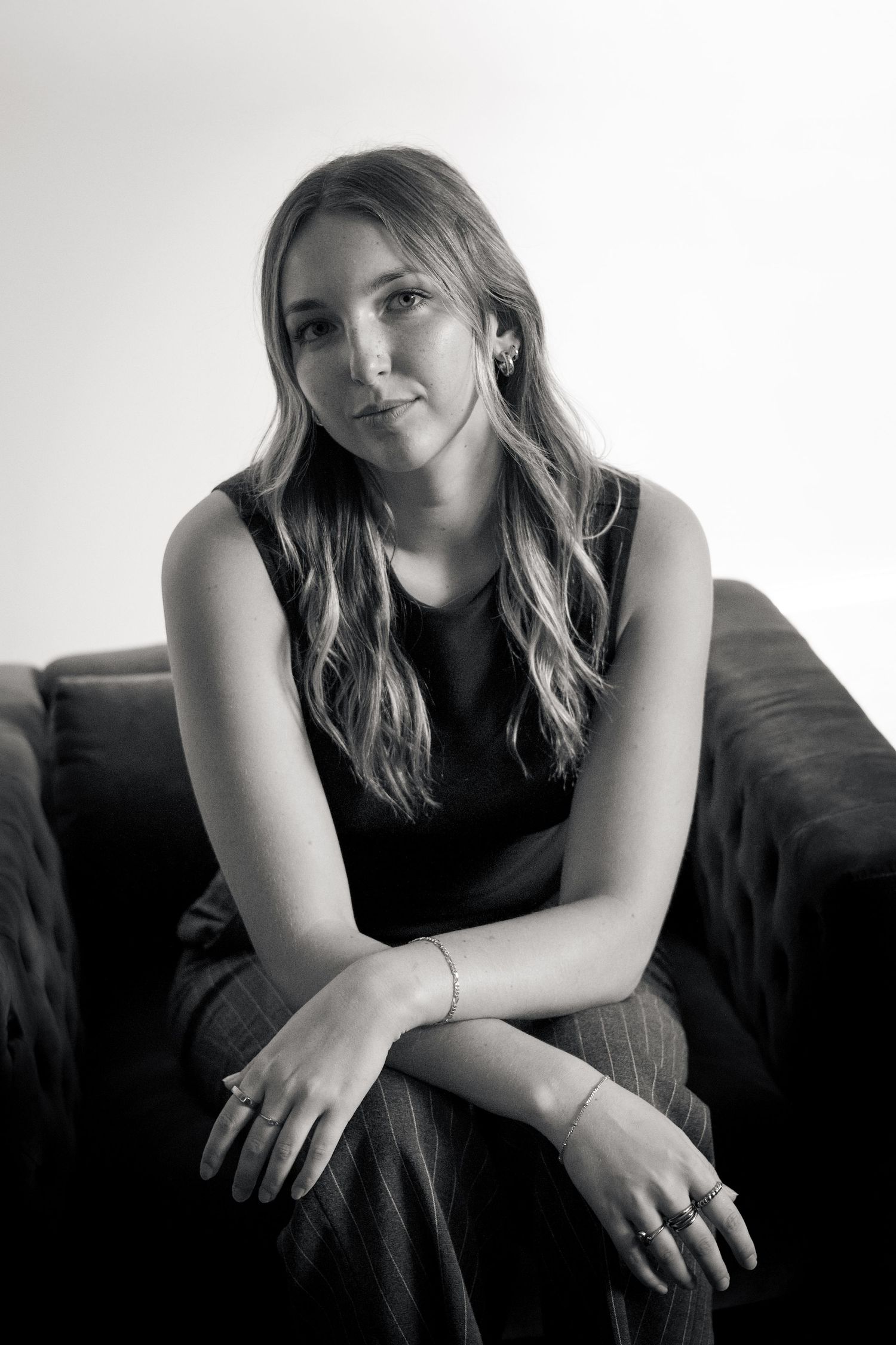 Black and white portrait of a young woman with wavy hair sitting on a sofa, wearing a sleeveless top.