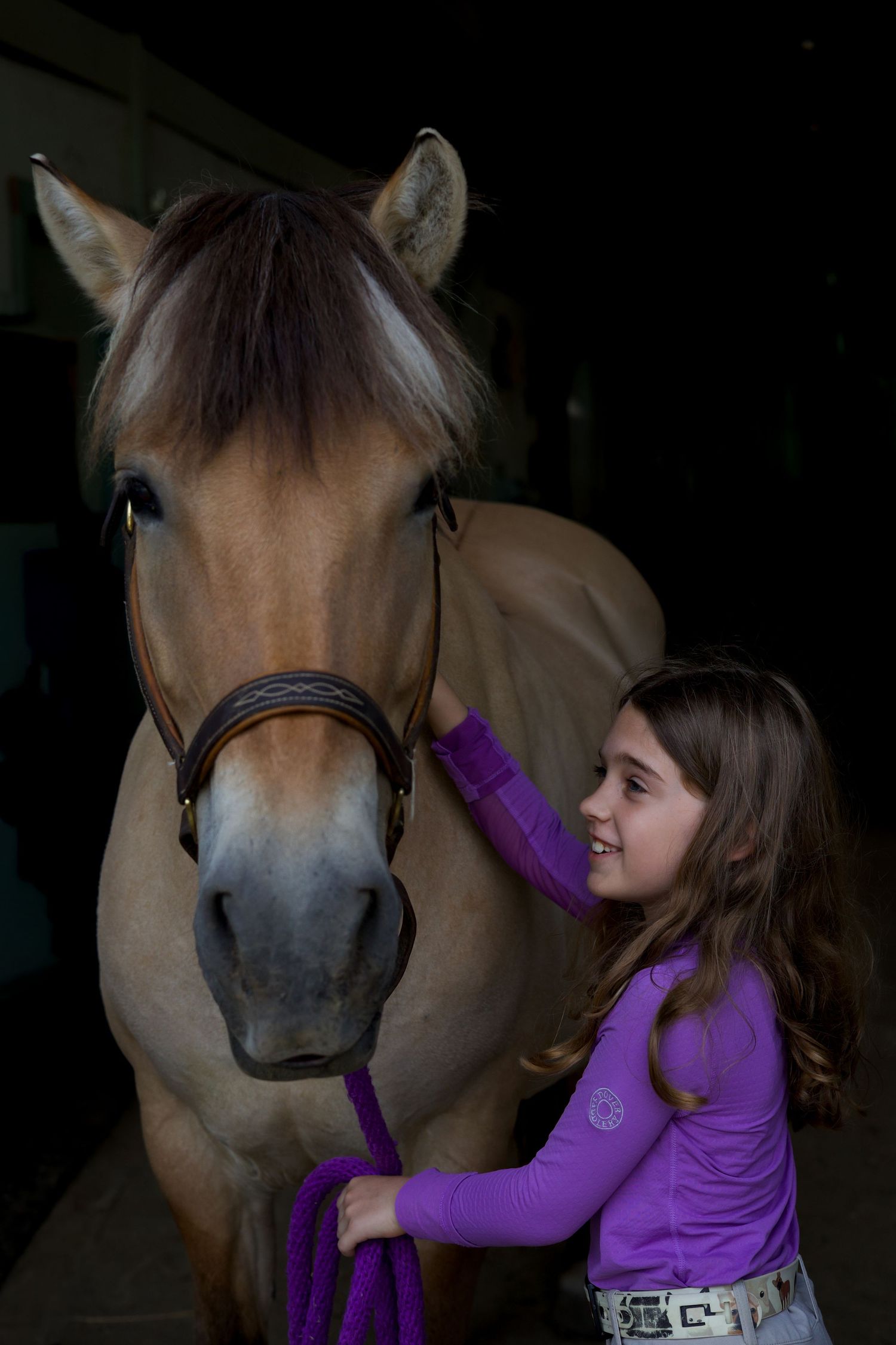 Smiling young girl in purple shirt gently petting a brown horse wearing a bridle in a dark stable.