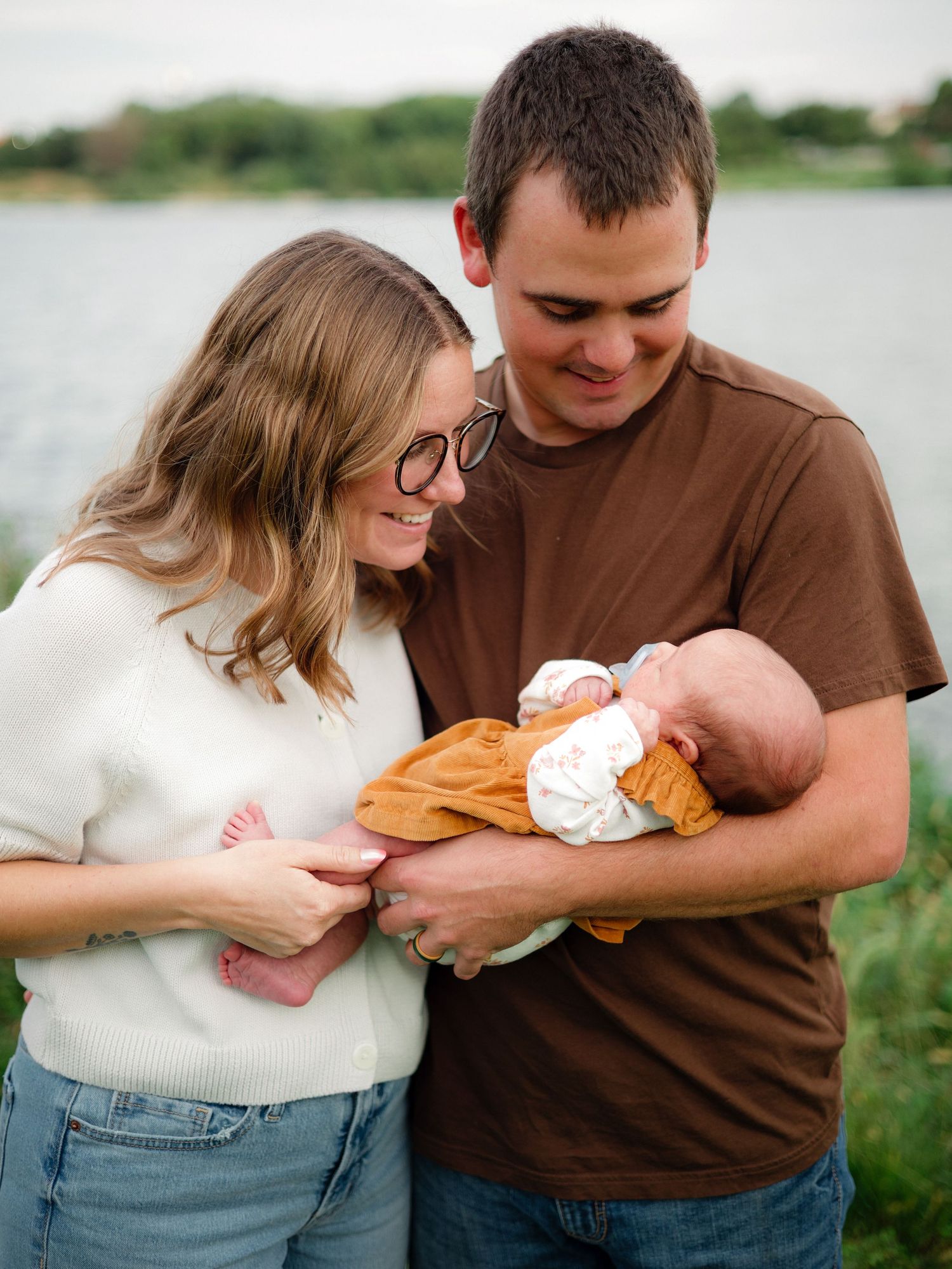 Young couple smiling at their newborn baby outdoors near a lake, family portrait photography.