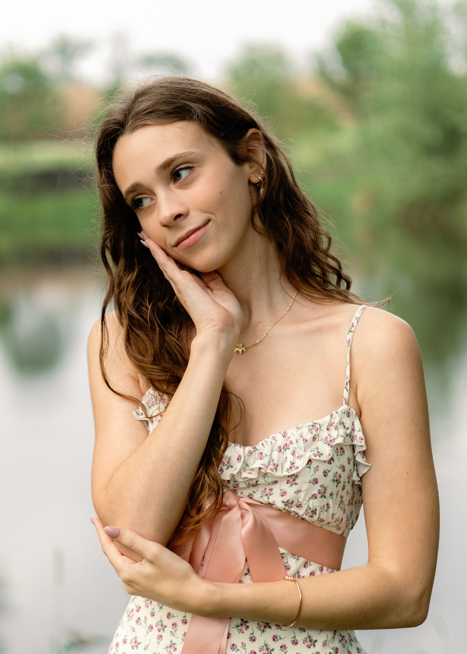 Young woman in floral dress posing thoughtfully by a lake, hand on cheek, with soft green bokeh background.