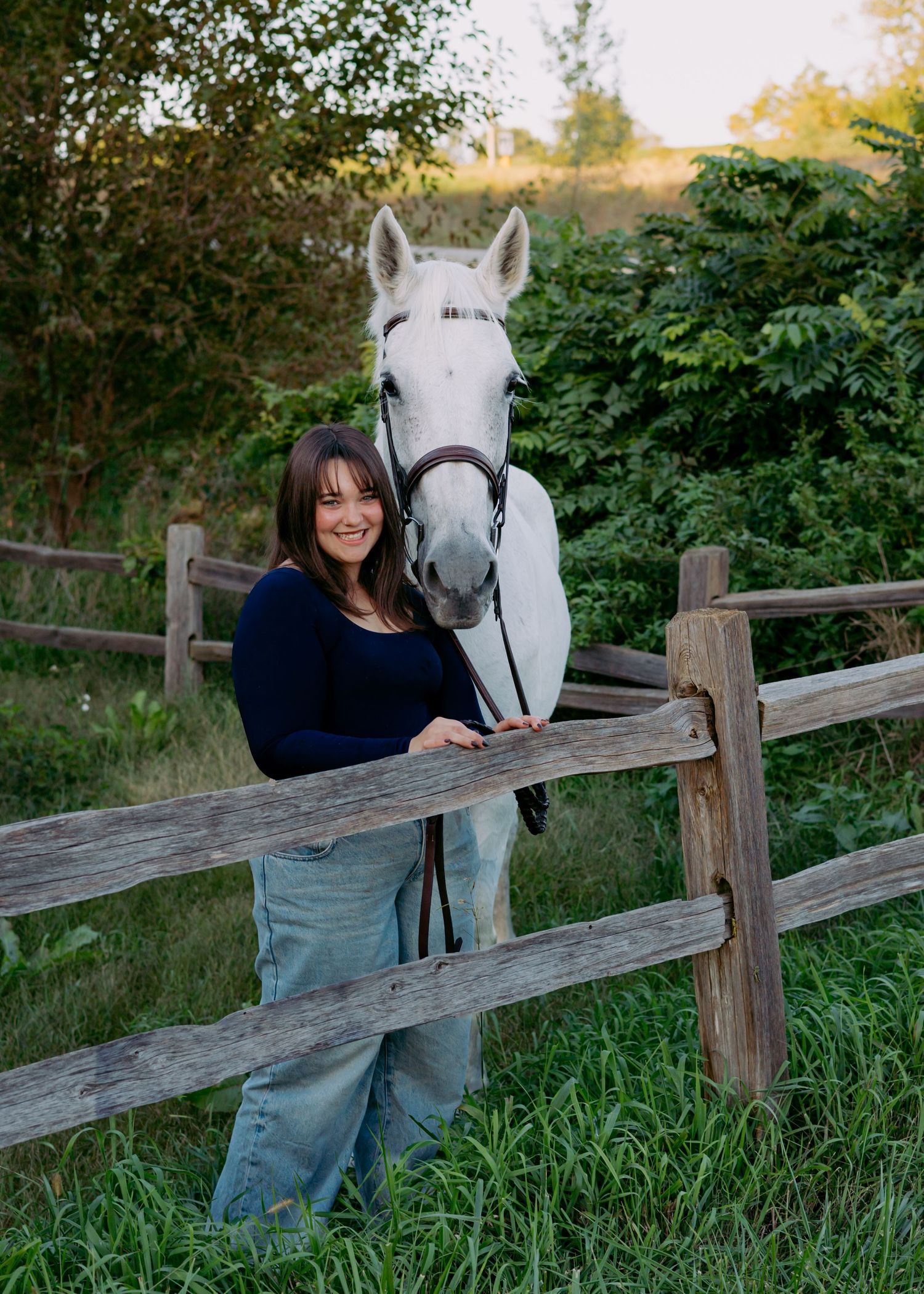 Smiling woman in a navy top and jeans stands beside a white horse in a bridle behind a rustic wooden fence in a grassy green pasture.