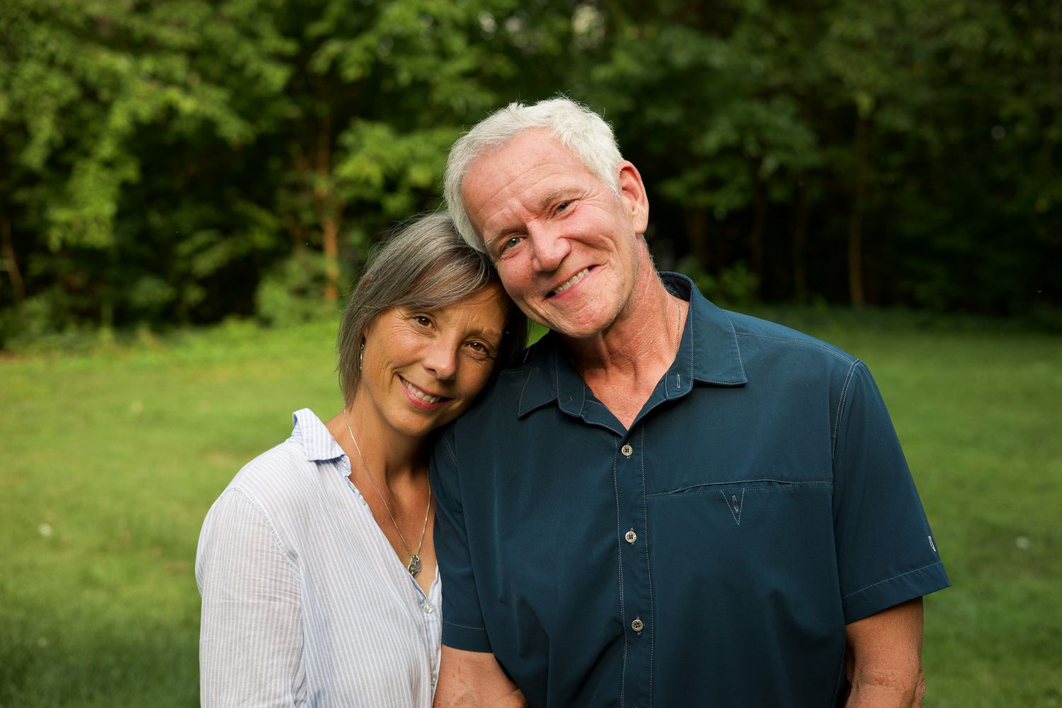 Smiling older couple embracing outdoors on a green lawn with lush trees behind them, the man in a dark blue shirt and the woman in white.