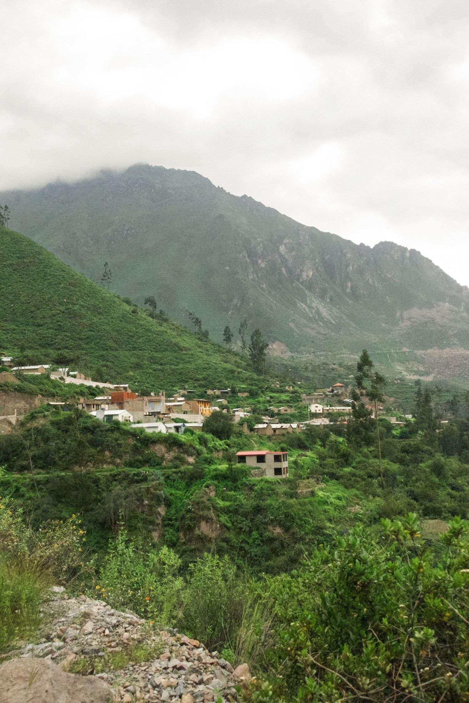 A small Andean village nestled among lush green hills with a dramatic mountain peak shrouded in clouds.