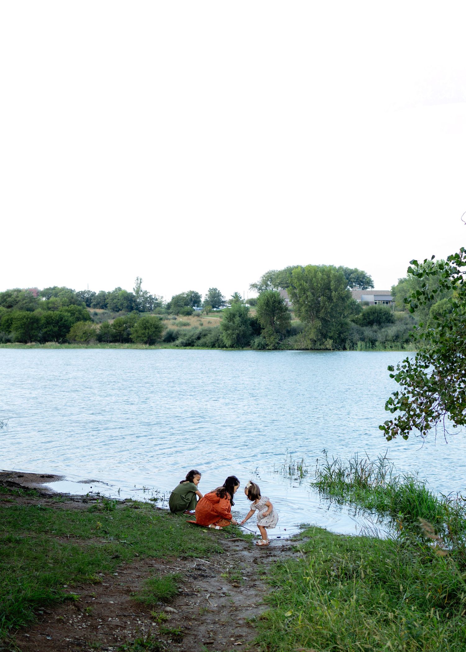 Family sitting by a calm lake shore surrounded by lush green trees on a cloudy day.