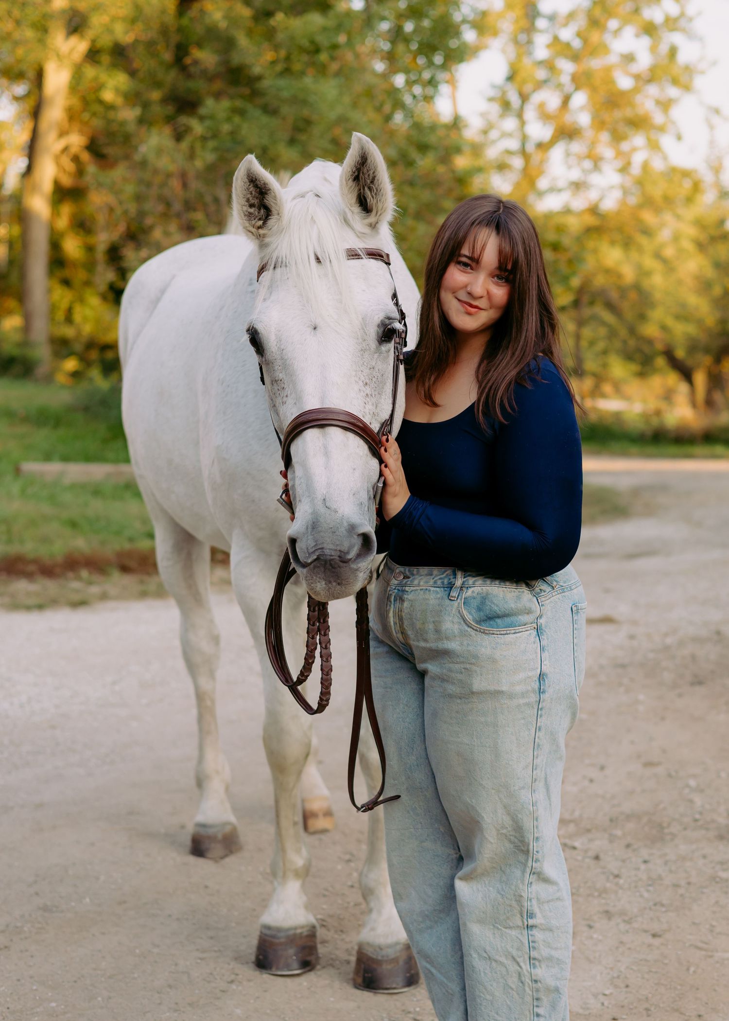 Smiling young woman in a navy sweater and light jeans stands beside a white horse wearing a leather bridle on a gravel path.