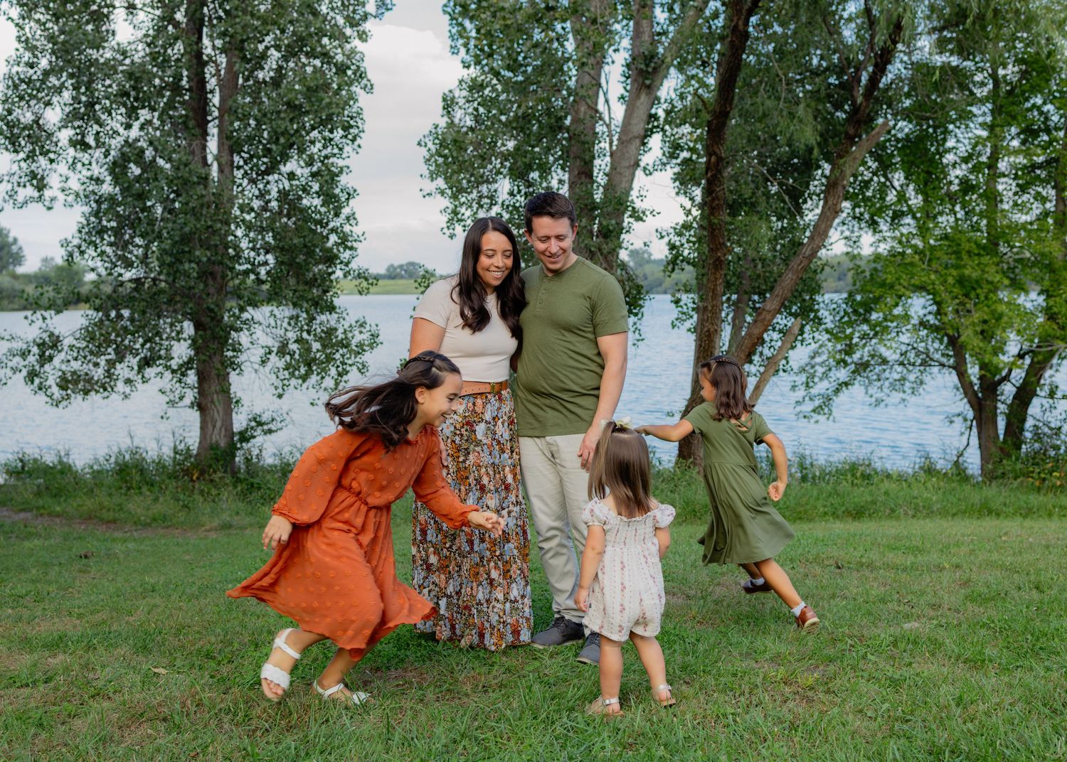 A smiling couple stands on a grassy lakeside lawn while three young girls in dresses play and twirl around them under tall trees.