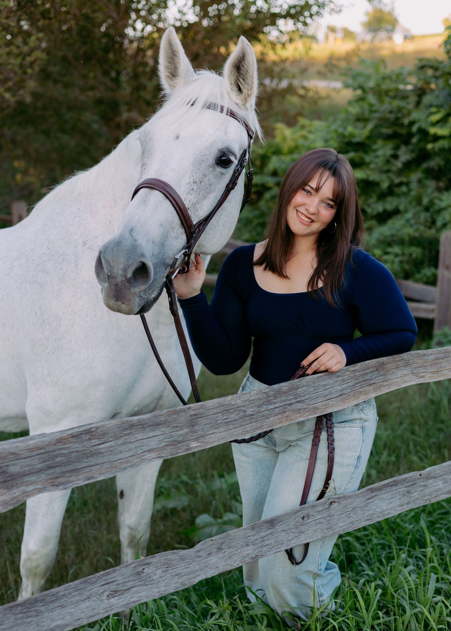 Smiling woman in a navy sweater stands beside a gray horse in a bridle at a wooden fence on a sunny green pasture.